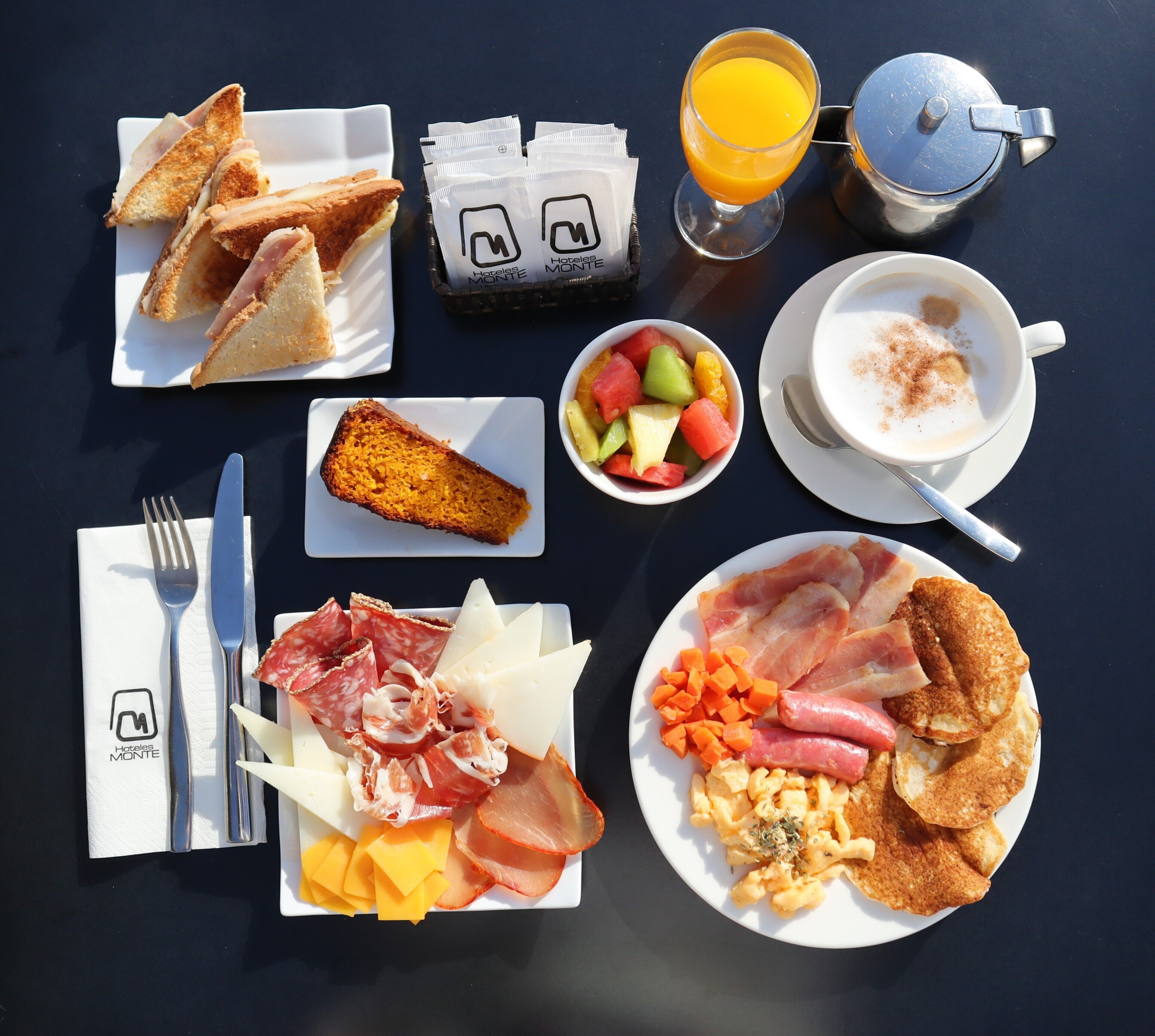 Une table de petit-déjeuner avec des tartines, des fruits, des œufs, du bacon, du fromage et une cafetière.