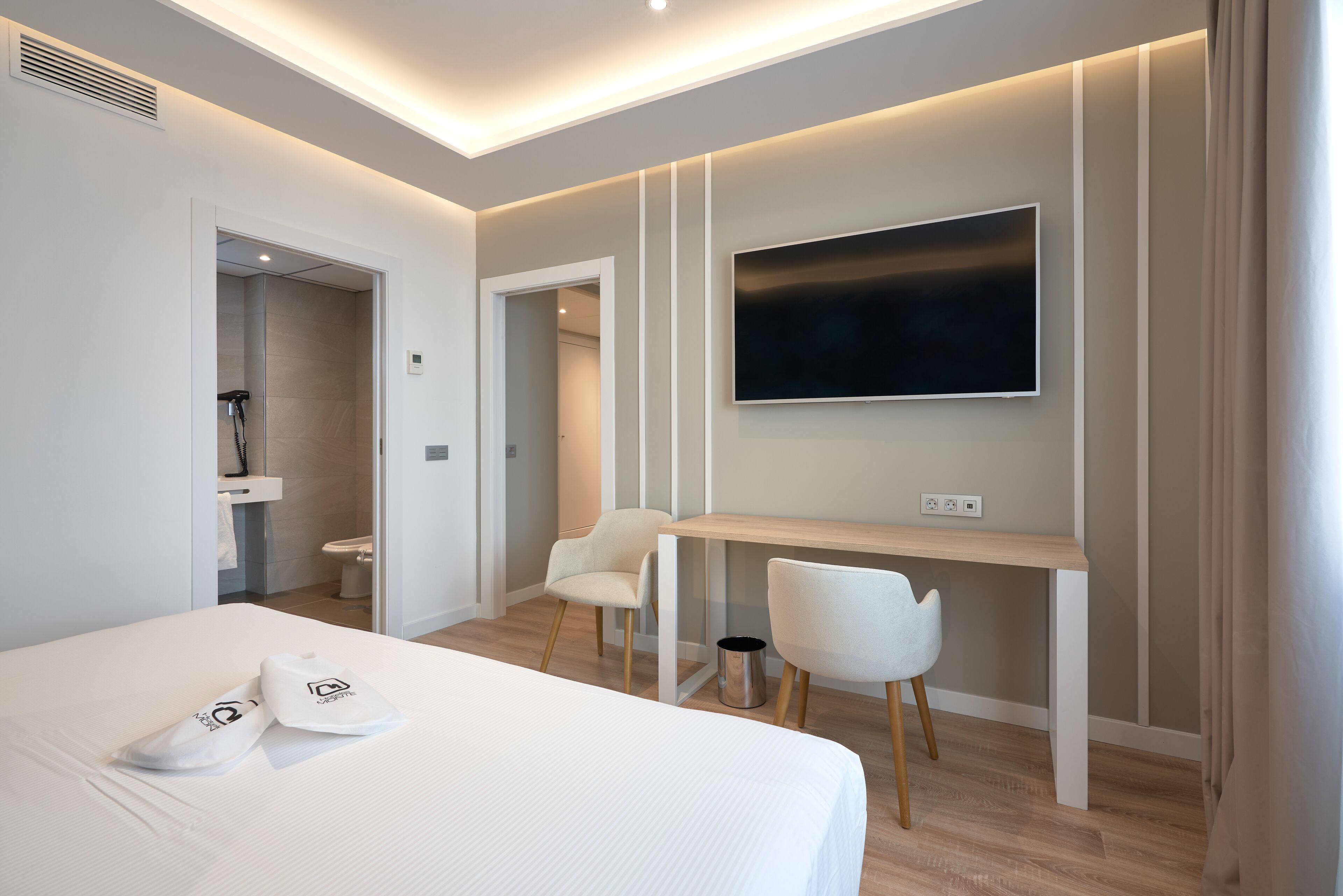 A modern hotel room with a bed, a desk, two chairs, a TV, and a shower area.