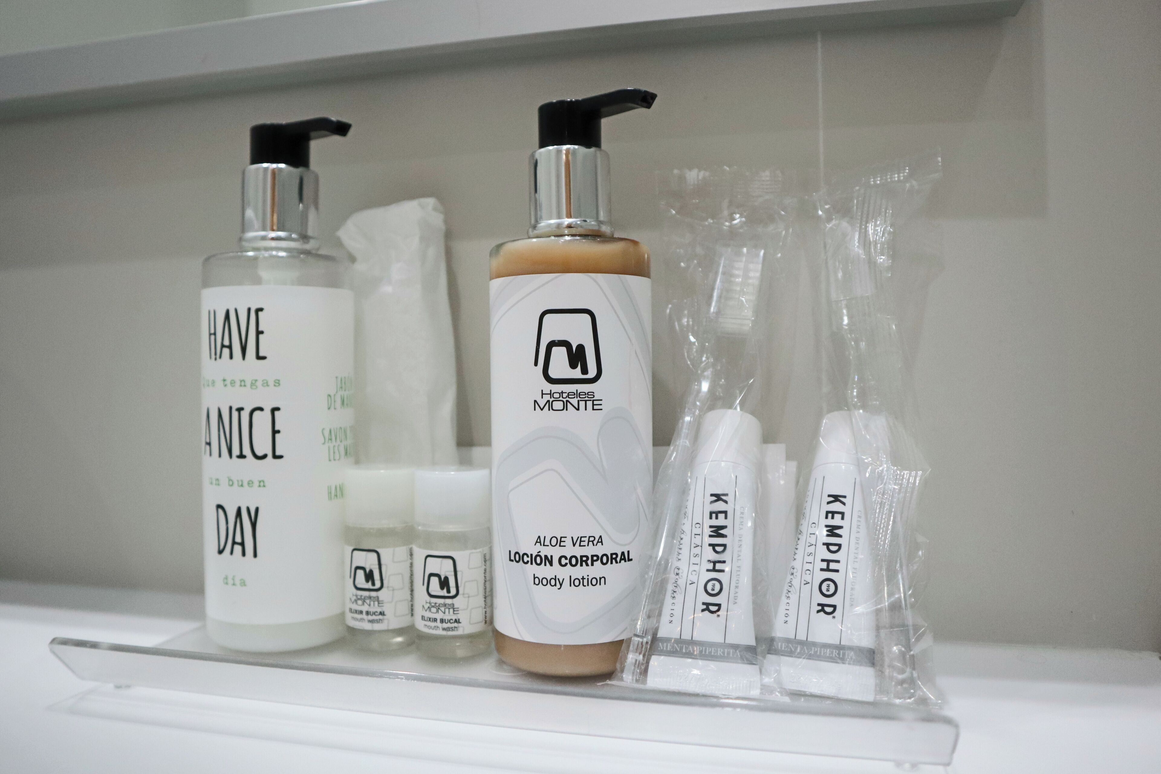 Una repisa de baño con diversos artículos de tocador, incluyendo una botella etiquetada 'HAVE ANICE DAY', una botella etiquetada 'Aloe Vera Lloción Corporal' y varios otros productos de cuidado personal.