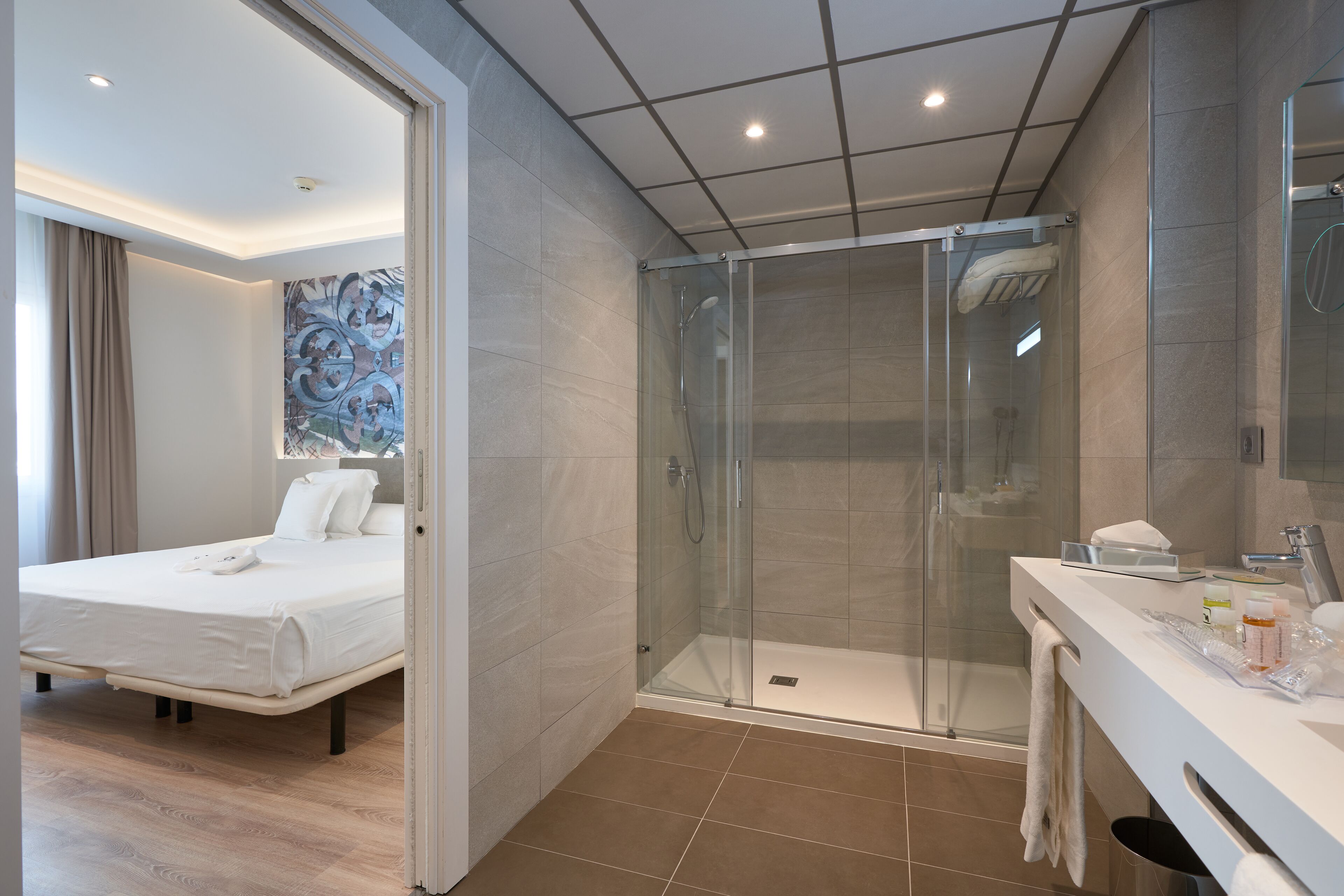 Una habitación de hotel moderna con una cama, ducha acristalada y baño con tocador y espejo.