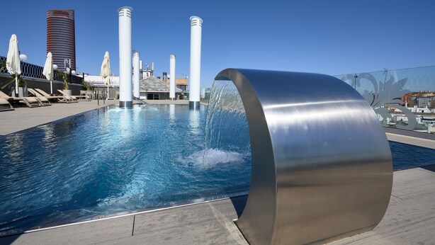 Una piscina con fuente de agua, sillas de descanso y arquitectura moderna al fondo.
