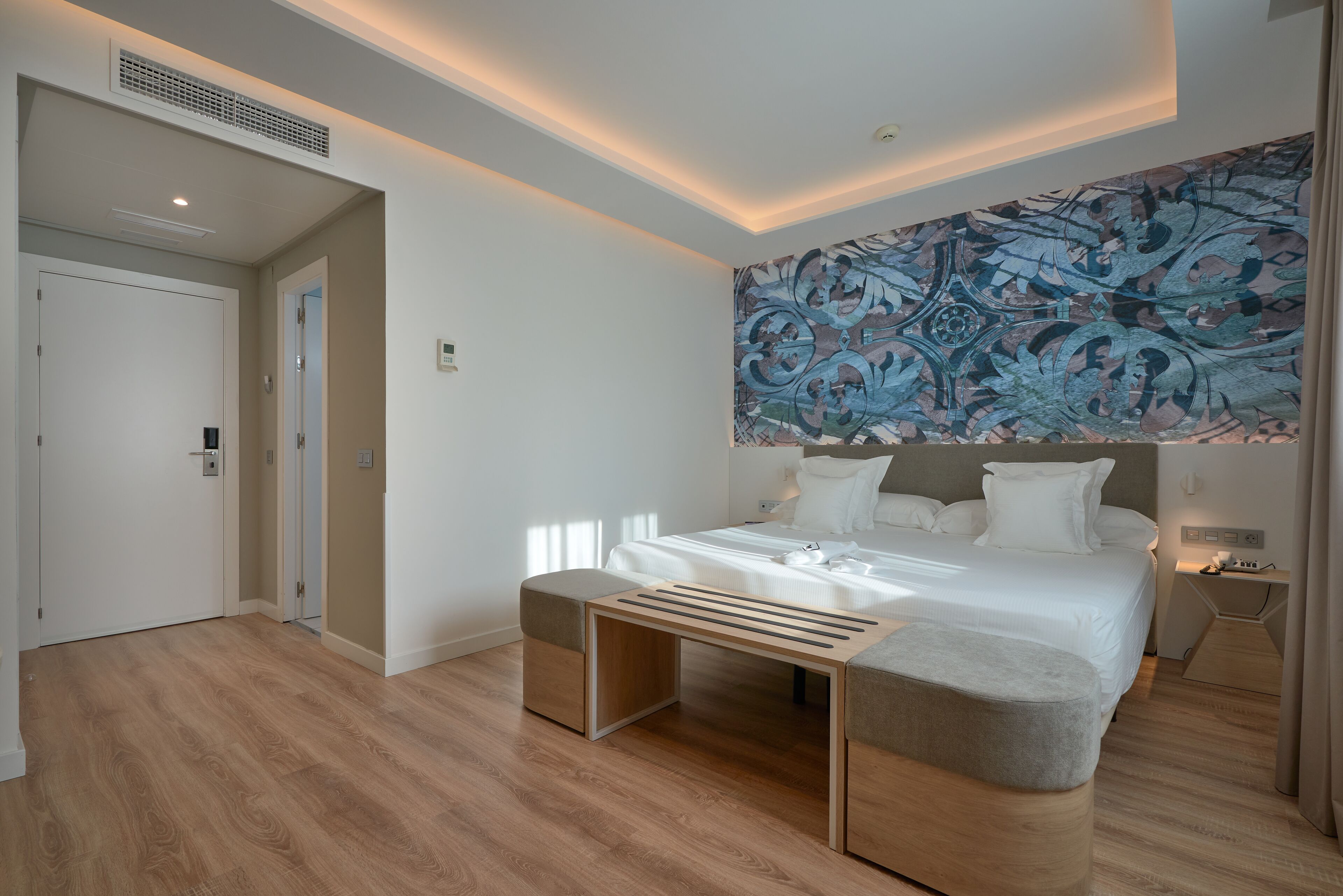 A modern hotel room with a large bed, a bench, a wall-mounted artwork, and a door.