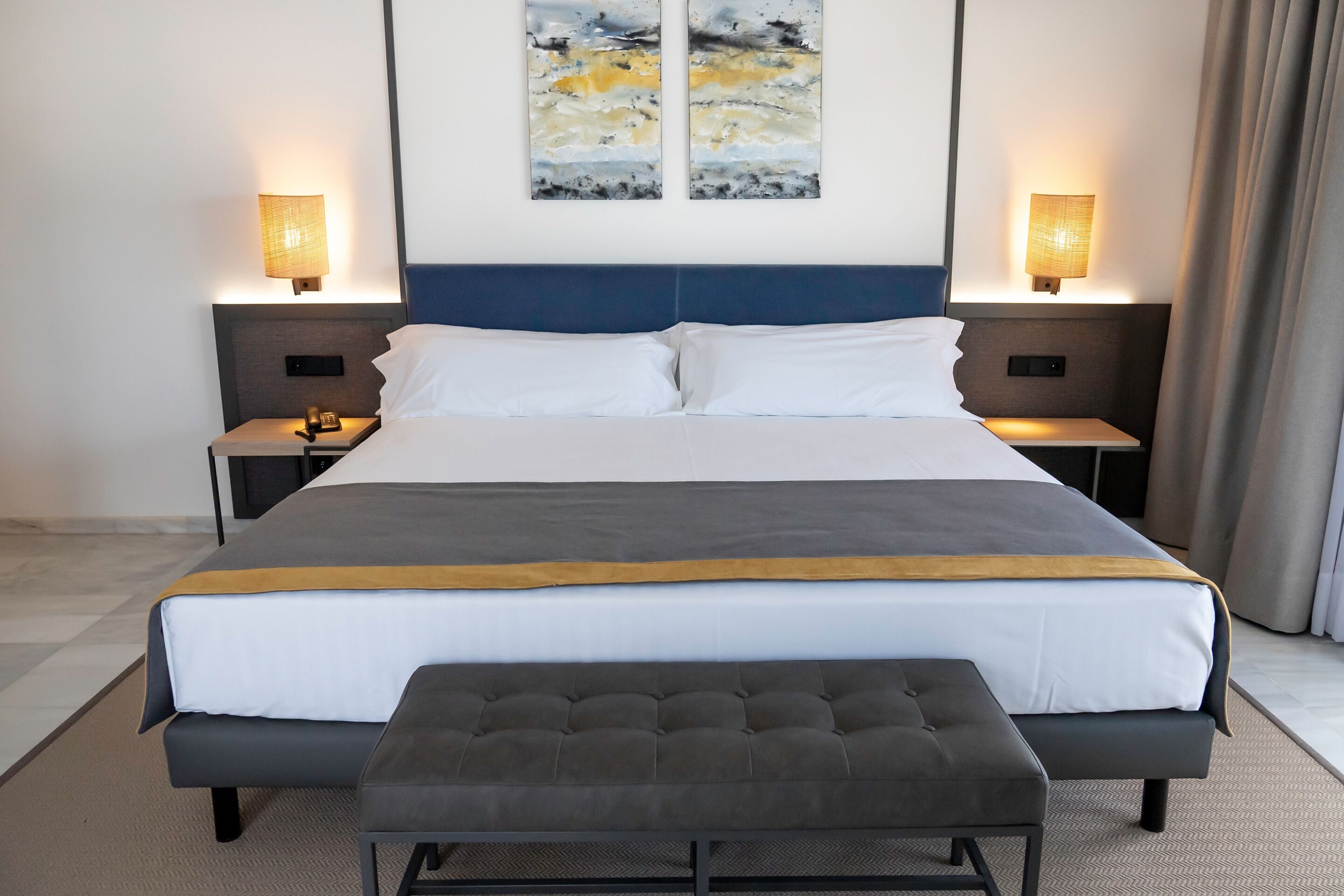 Une chambre d’hôtel moderne avec un grand lit, deux tables de chevet, un banc et des œuvres d’art abstraites accrochées au mur.