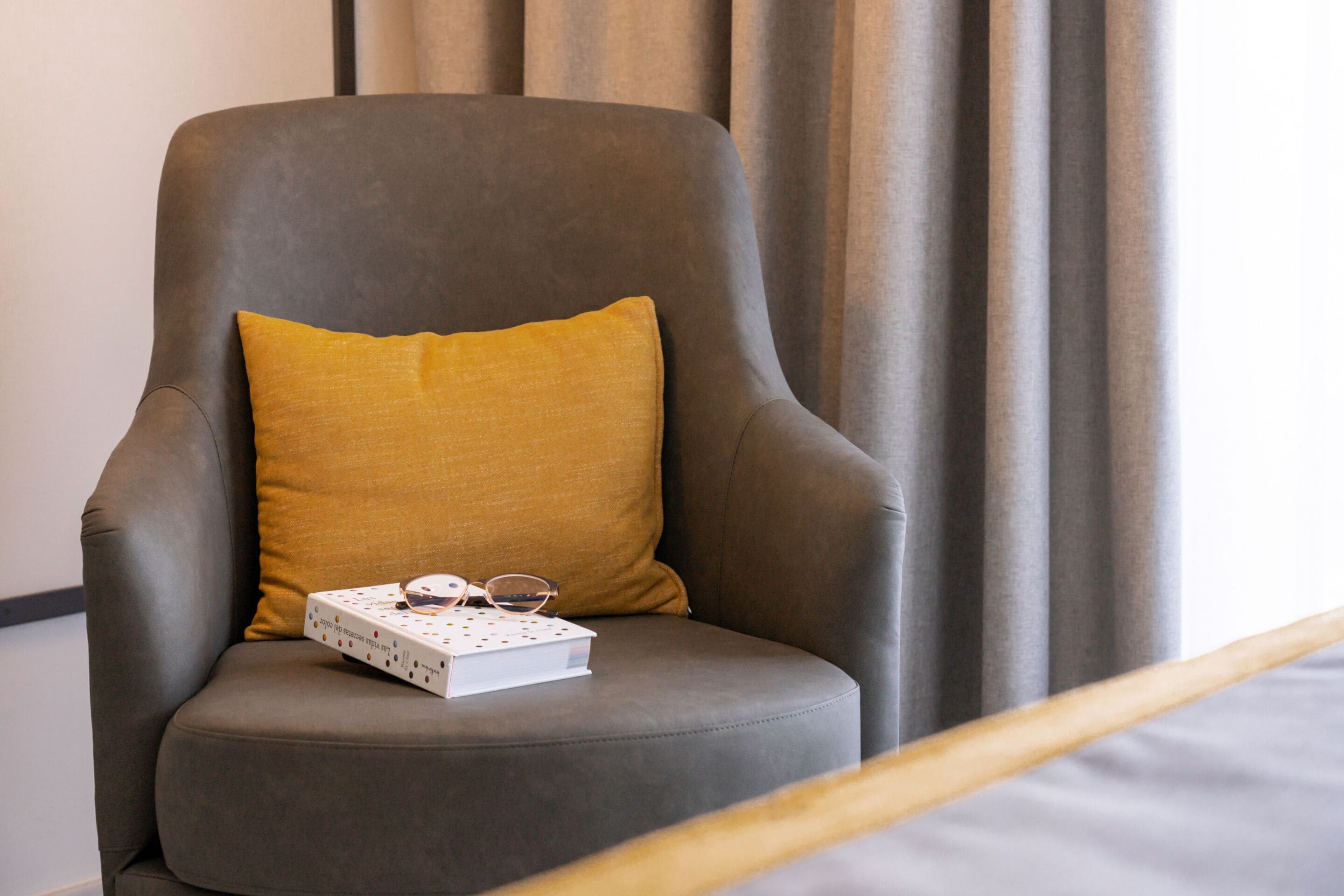 Un sillón gris con un cojín amarillo mostaza y una caja blanca con gafas encima.