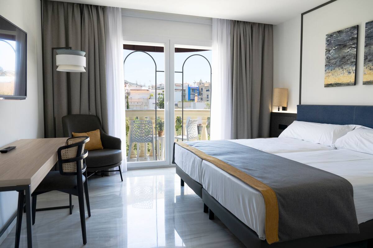 A hotel room with a bed, a desk, a chair, a balcony with curtains, and a view of buildings.