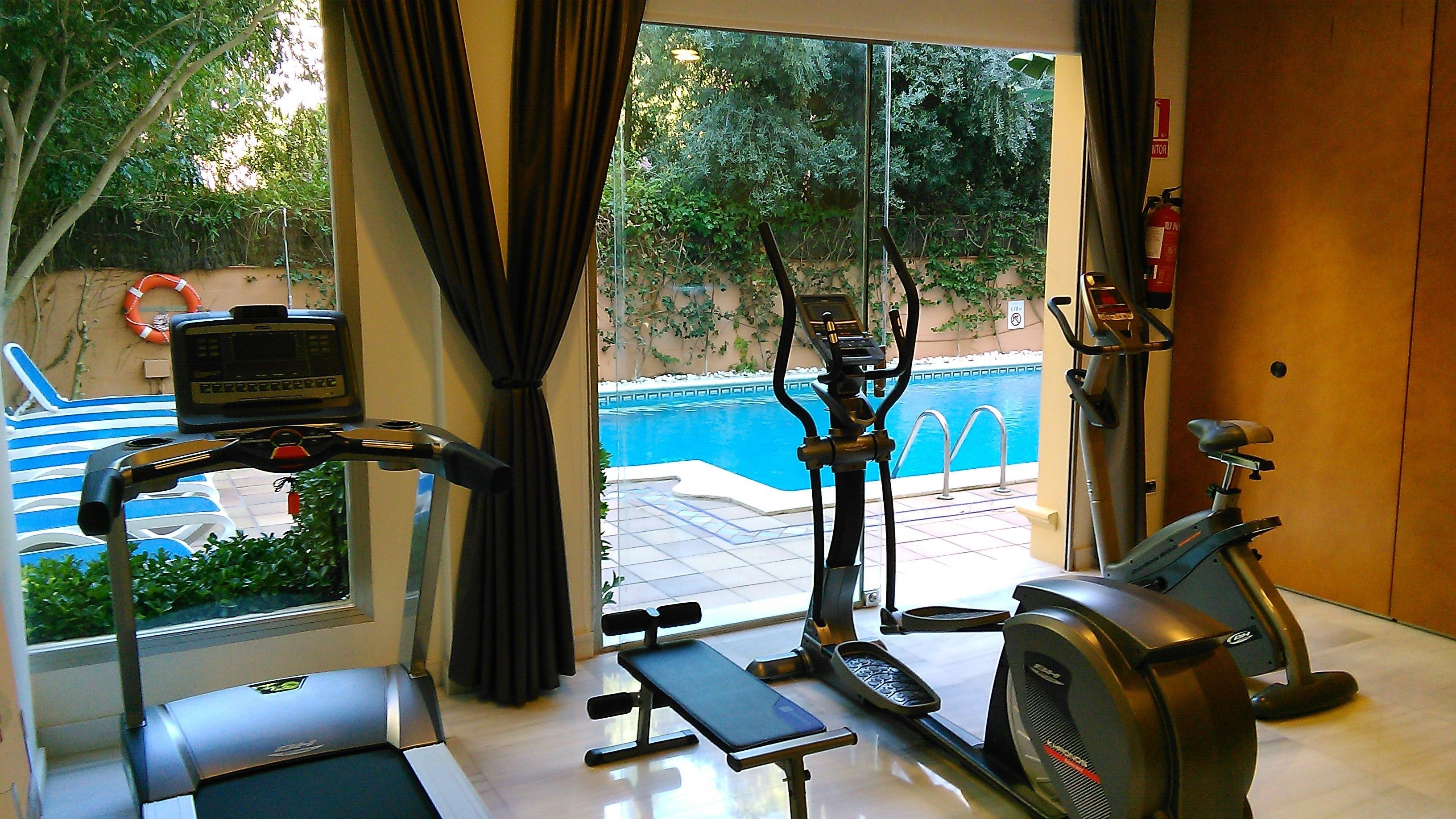 Un gimnasio en casa con cinta de correr, elíptica y banco, con vista a una piscina.