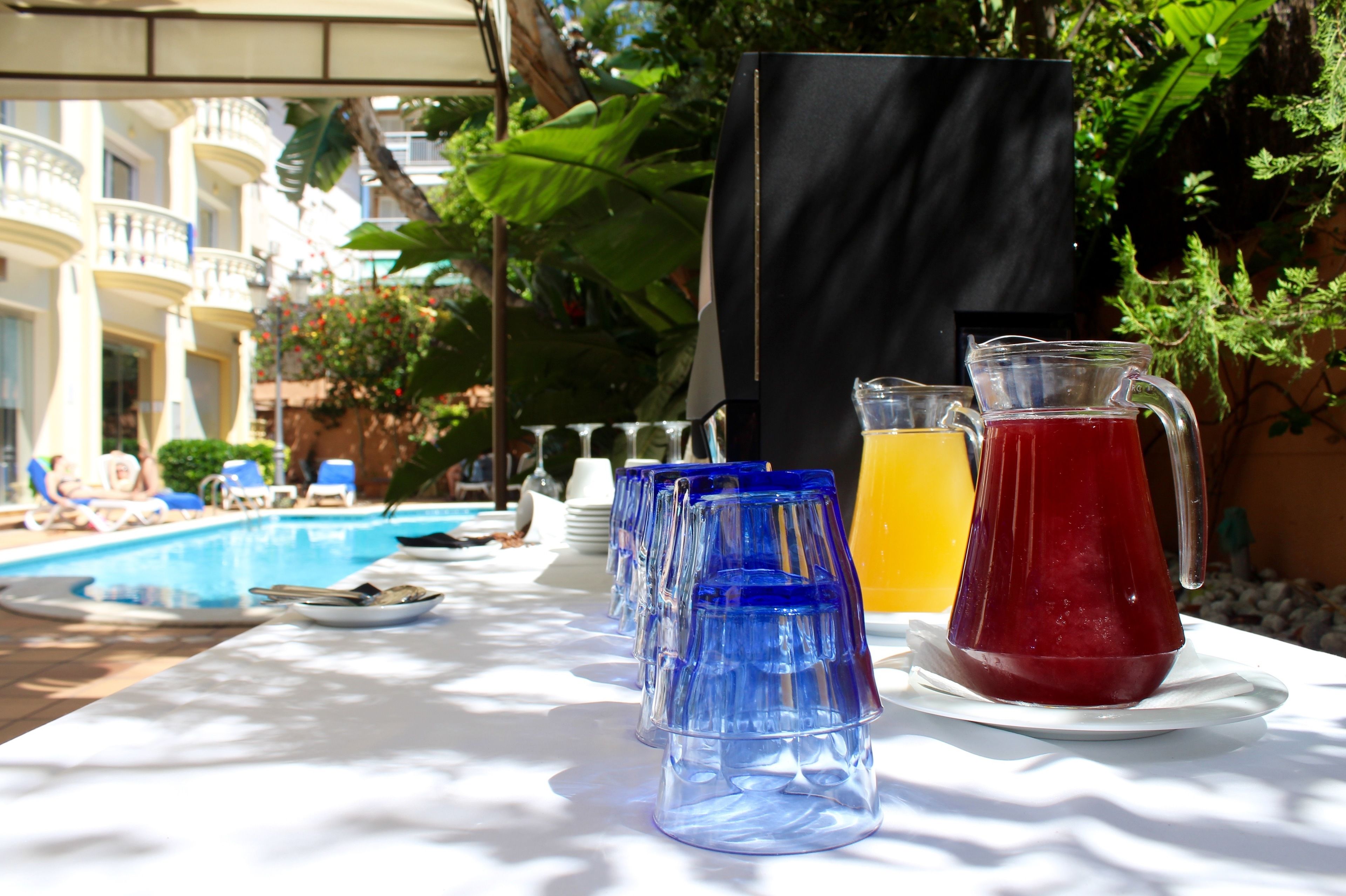 A table with blue glasses, a red pitcher, and a yellow drink, set outdoors with a pool and greenery in the background.