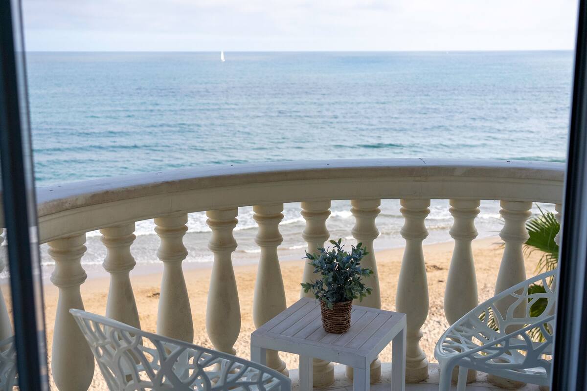 A balcony with a view of the sea, a small table with a plant, and white chairs.