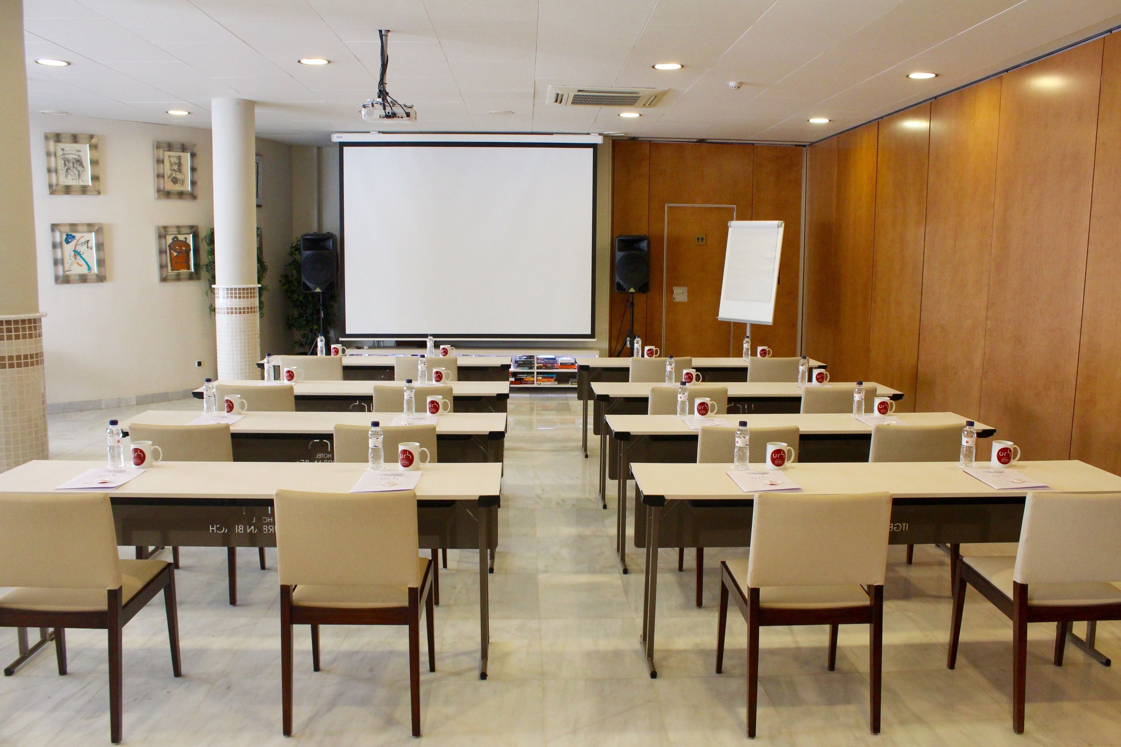A conference room with a large screen, multiple tables, and chairs, equipped with water bottles and name cards.
