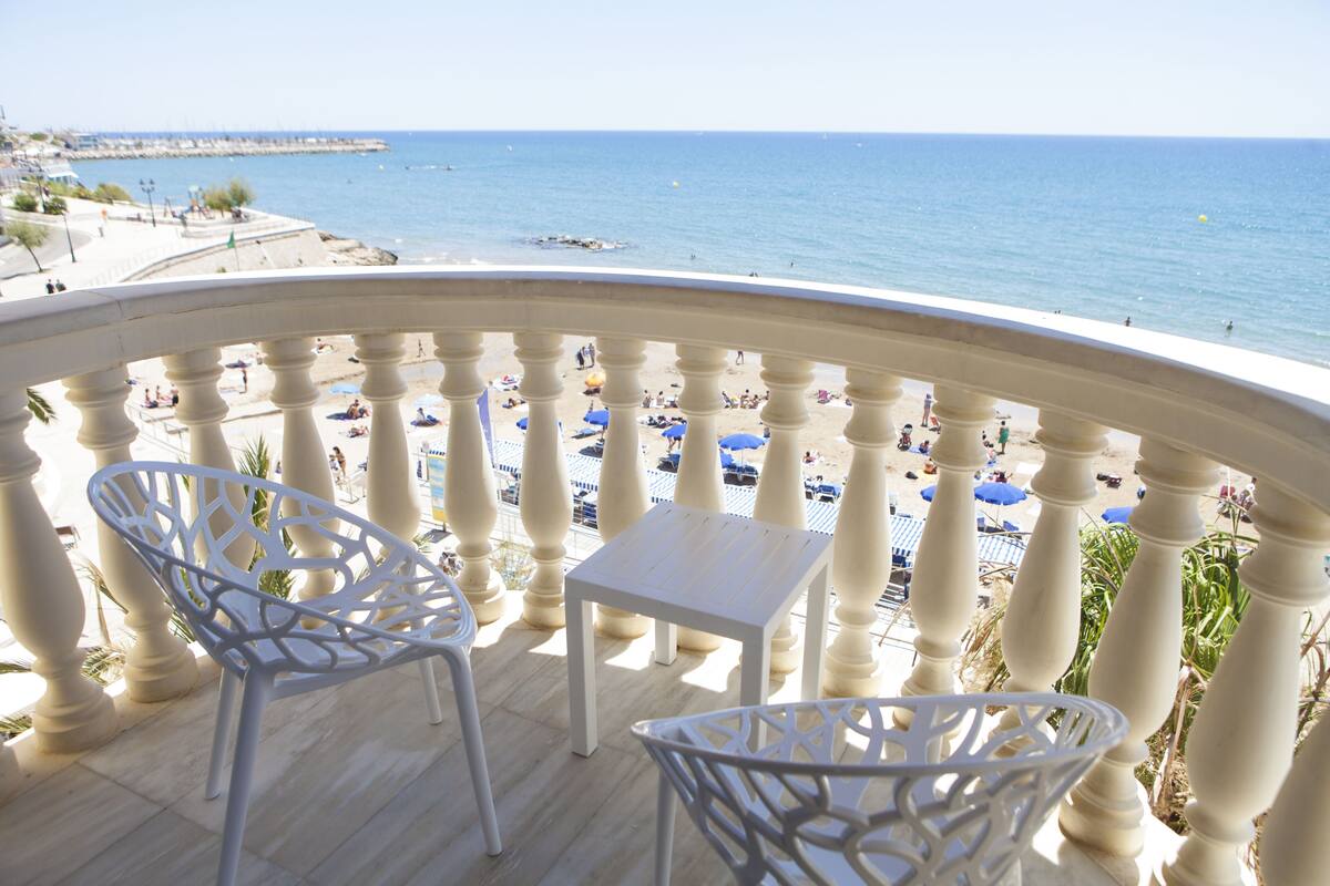 A balcony with white chairs and a small table, overlooking a beach with umbrellas and people.