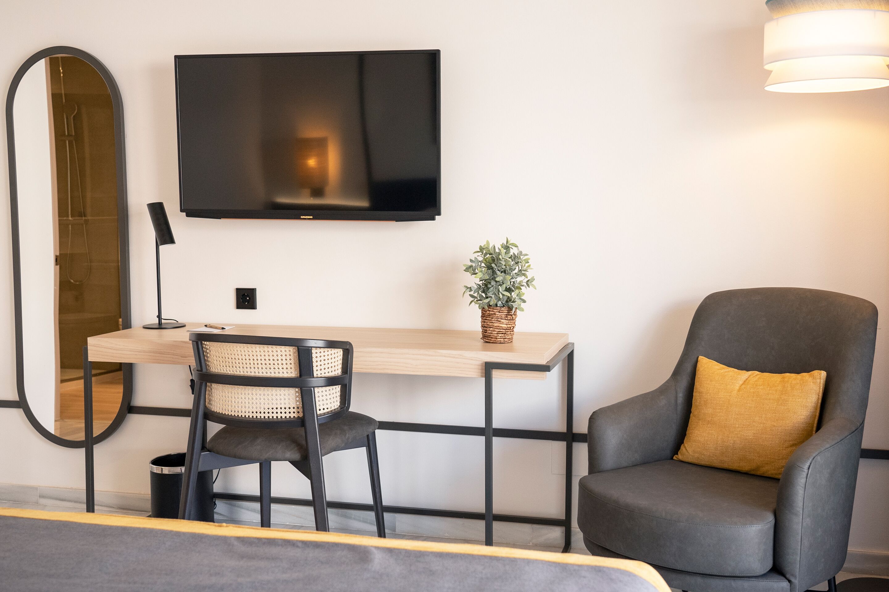 Een moderne hotelkamer met een houten bureau, een zwarte stoel, een grijze fauteuil met een geel kussen, een flatscreen televisie aan de muur en een kleine potplant.