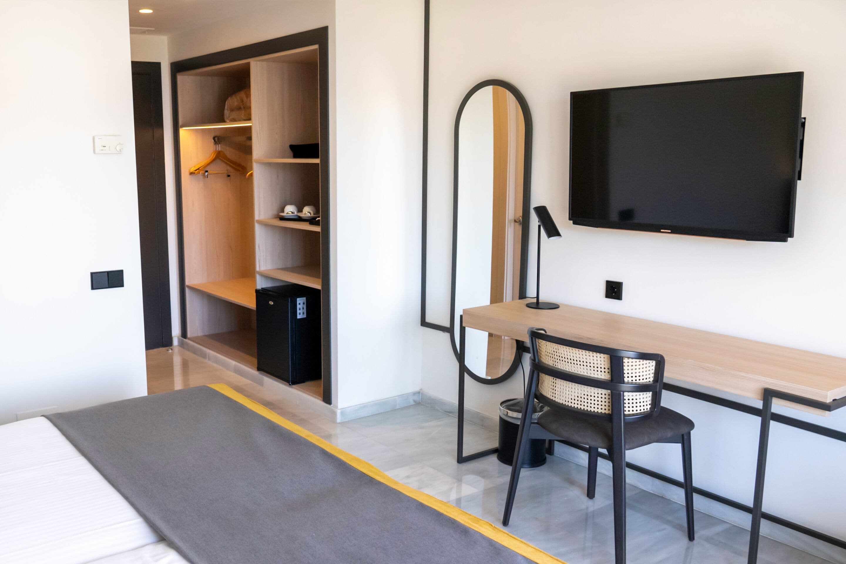 Una habitación de hotel moderna con cama, escritorio con silla, televisor, armario y espejo.