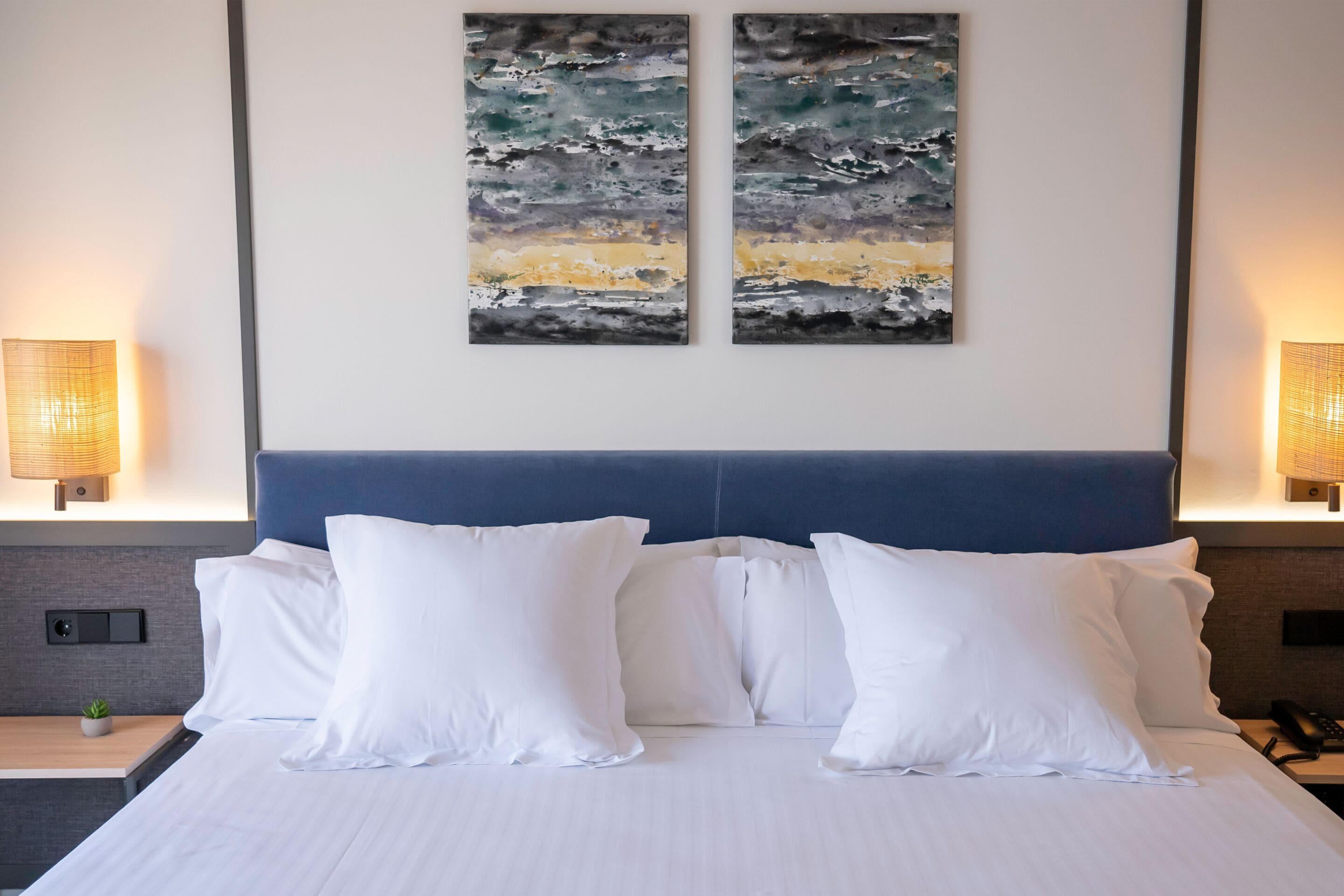 A hotel room with a large bed, white pillows, two abstract paintings on the wall, and bedside lamps.