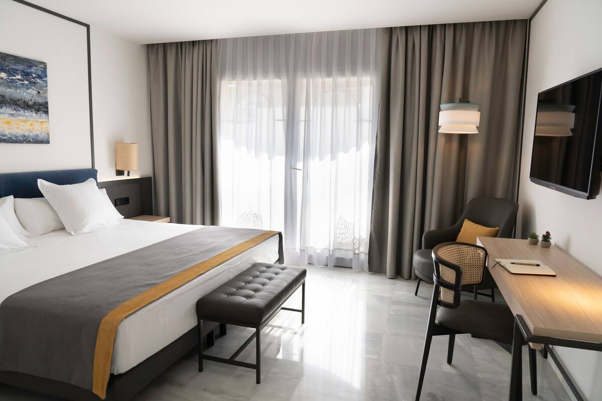 A modern hotel room with a large bed, a desk, a chair, and a view of the outdoors through sheer curtains.