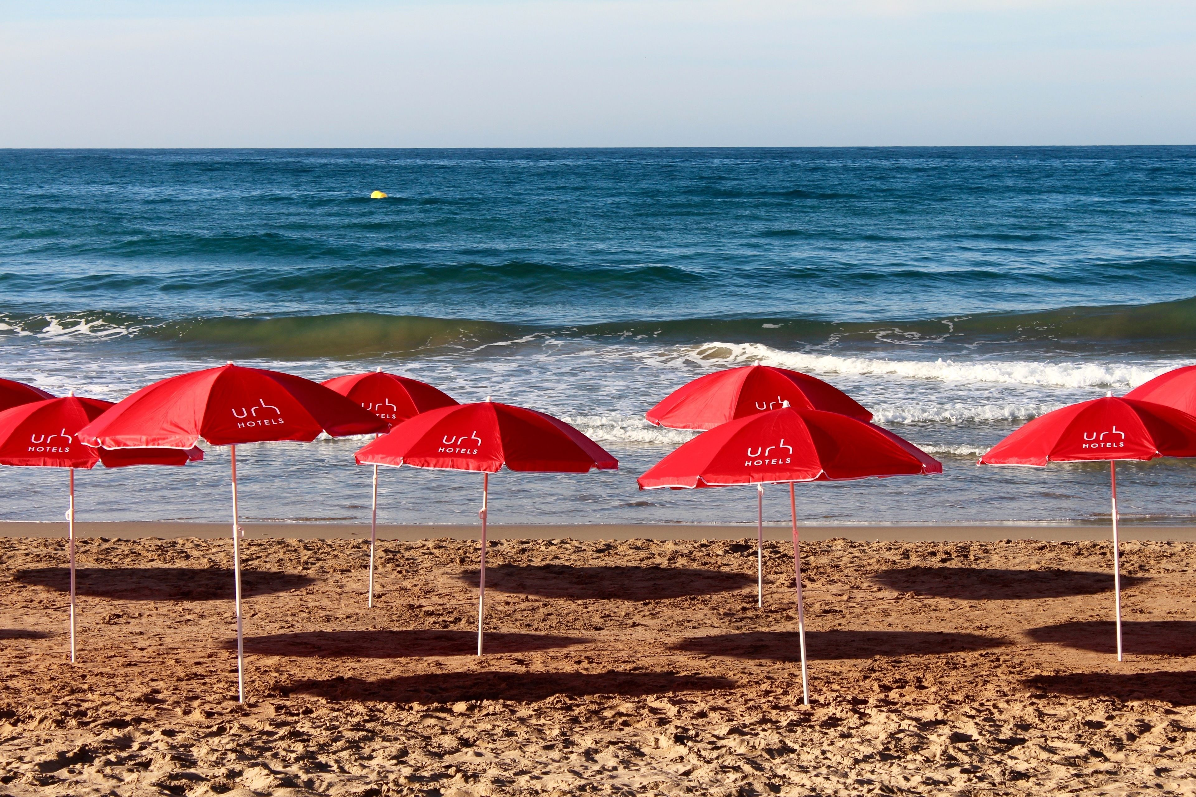 Une plage avec des parasols rouges sur lesquels on peut lire « URB HOTELS », et l’océan en arrière-plan.
