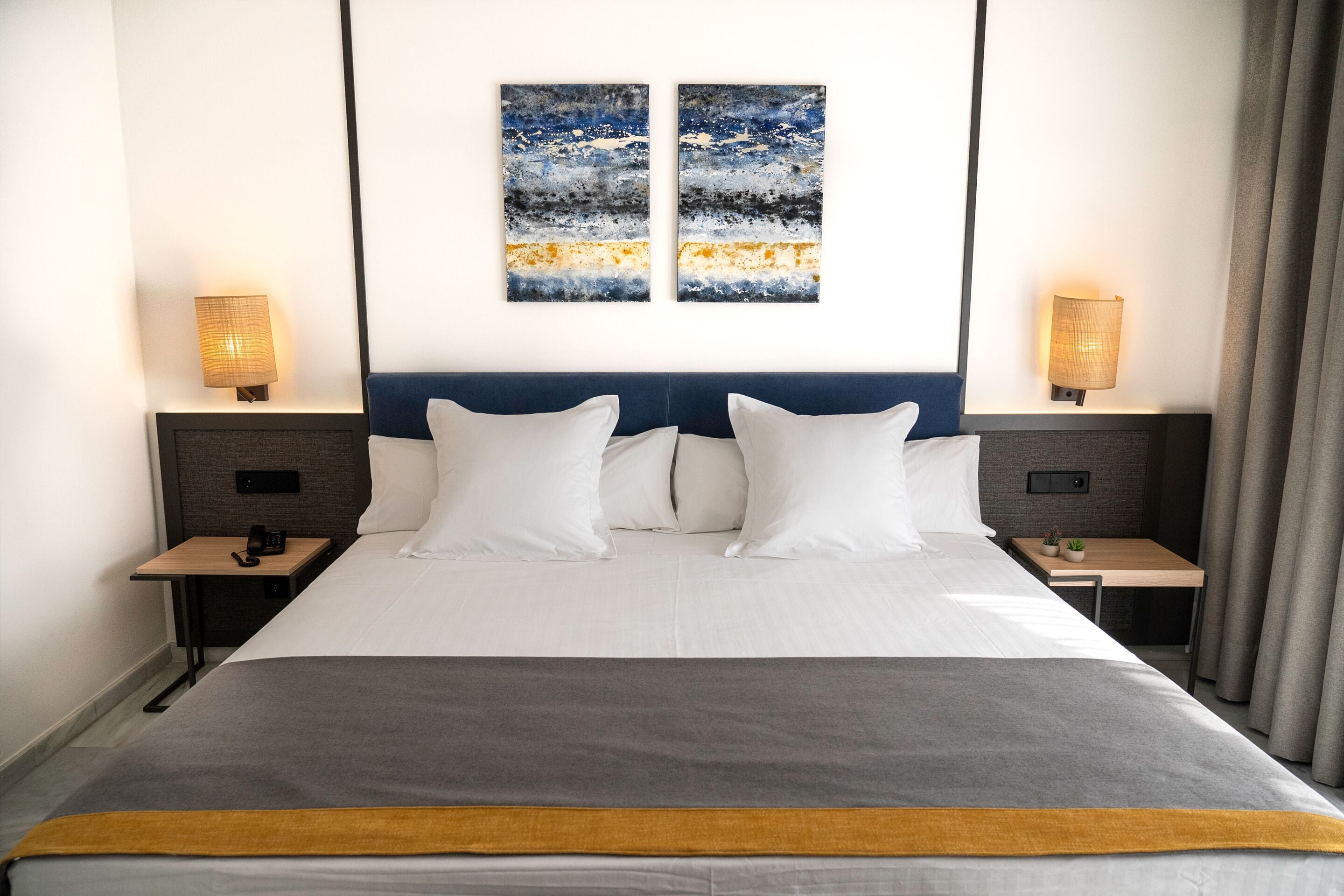 A modern hotel room with a large bed, two bedside tables, a nightstand, and a wall-mounted lamp.