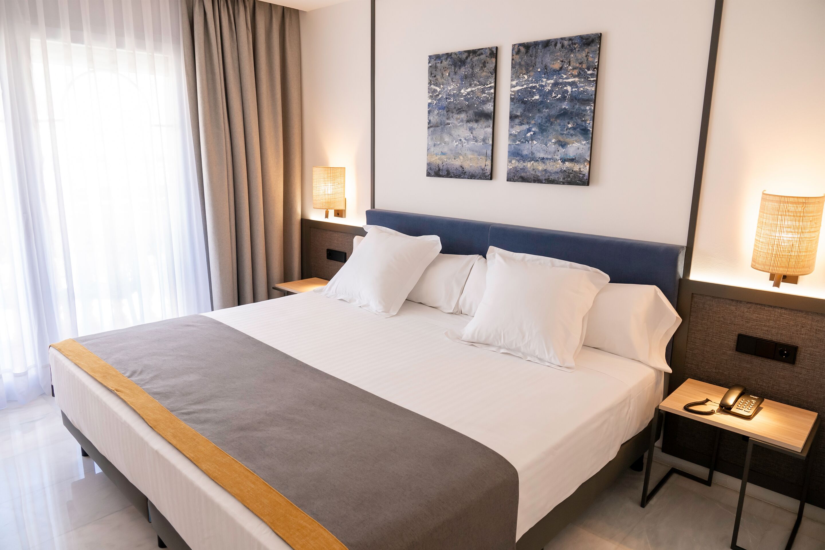 A hotel room with a large bed, two bedside tables, a lamp, and abstract artwork on the wall.