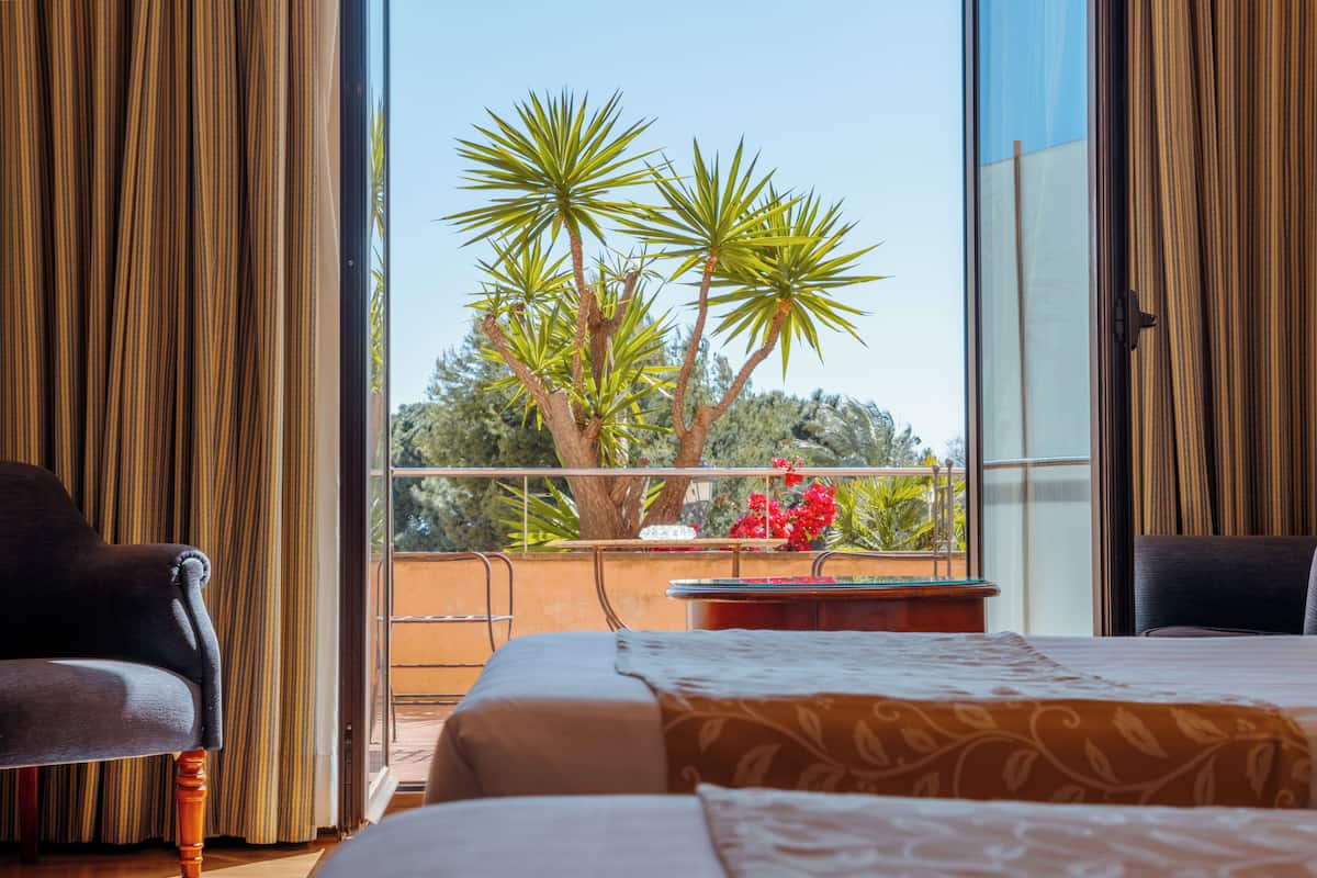A hotel room with a balcony, a bed, a chair, and a view of trees and flowers.