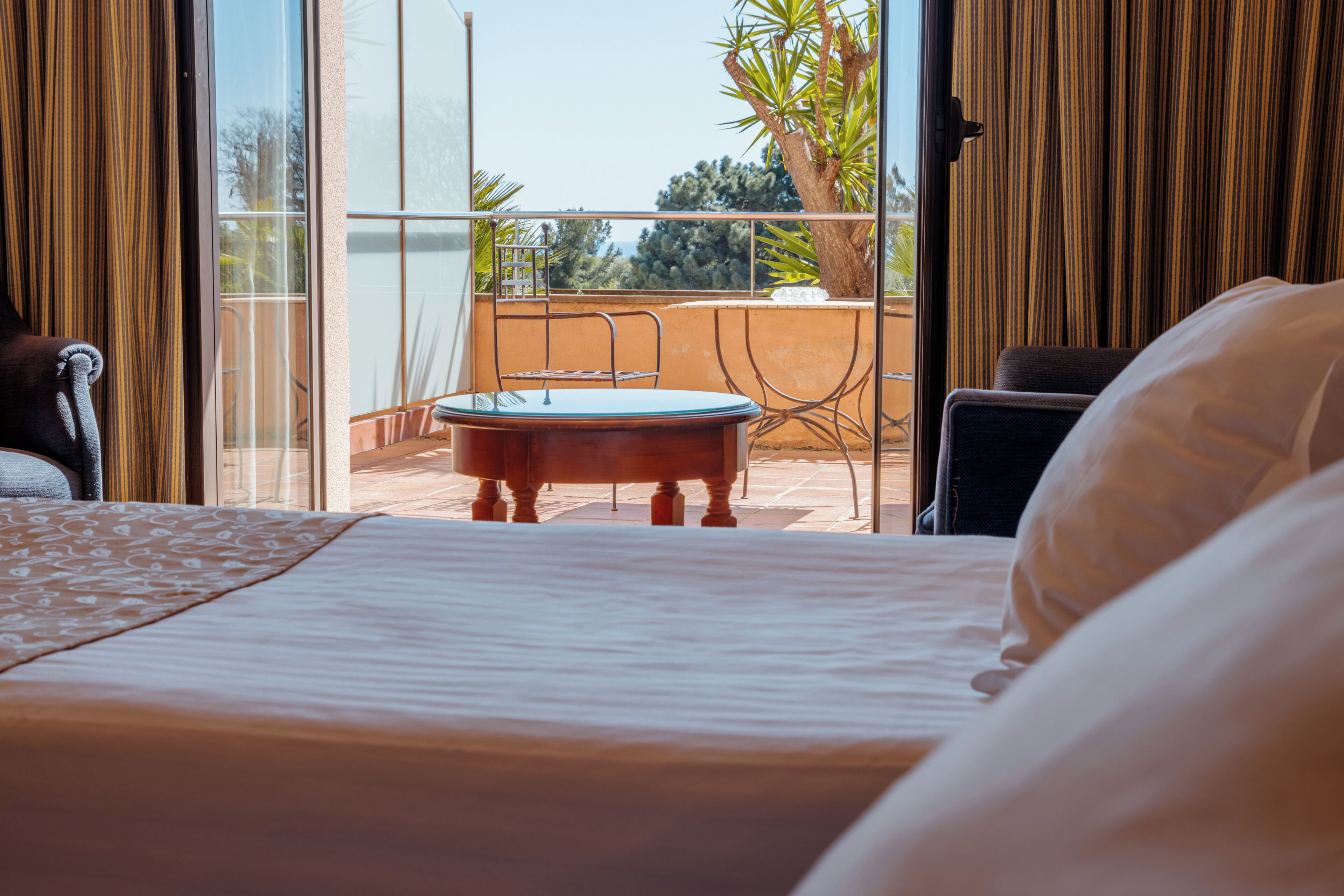 A hotel room with a bed, a small round table, and a view of the outdoors through a large window.