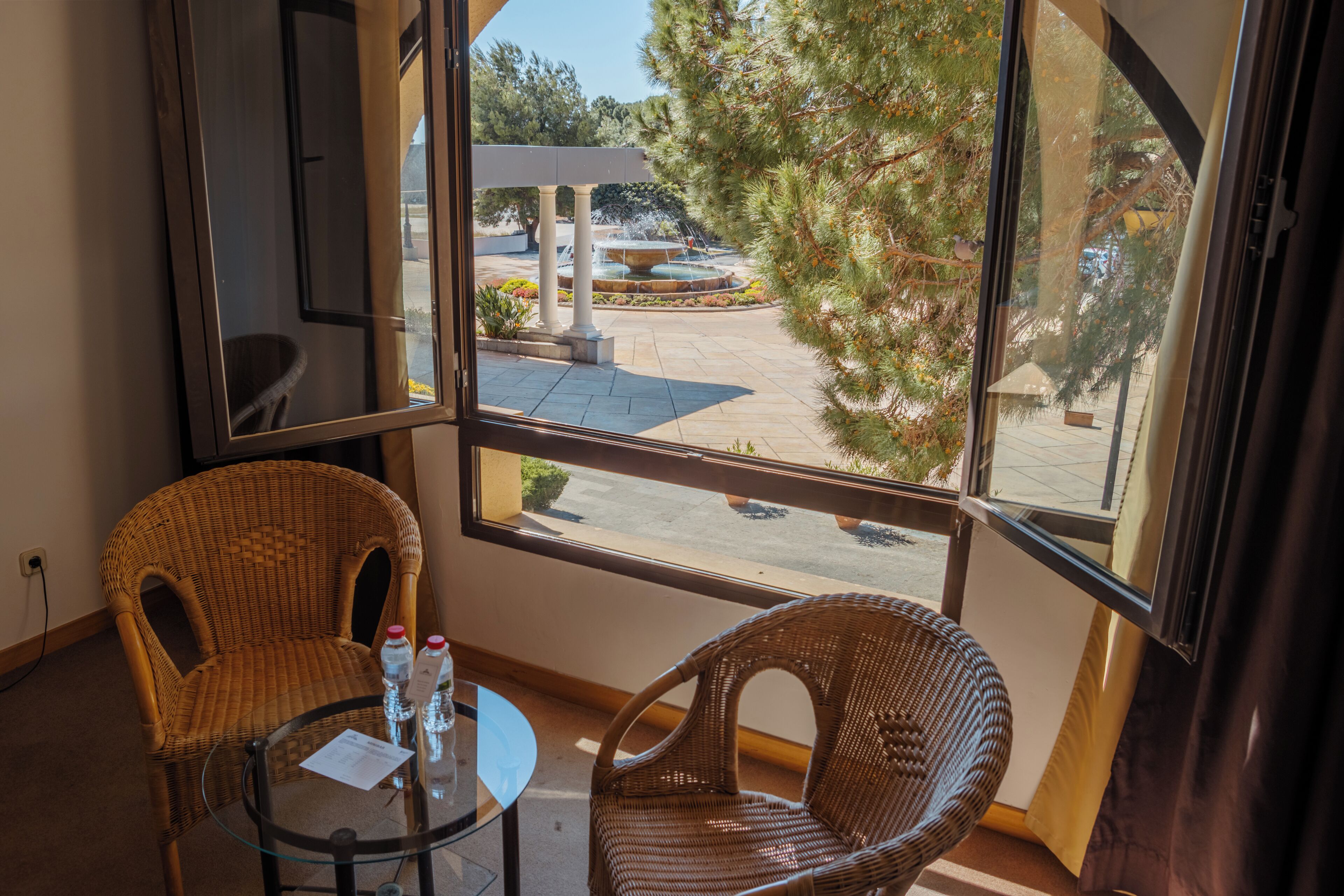 A room with two wicker chairs and a small table, a window with a view of an outdoor area including a fountain and trees.