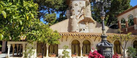 Un patio con un edificio histórico que presenta una torre, ventanas de arco y un tejado de tejas. Hay plantas en macetas y un farol.
