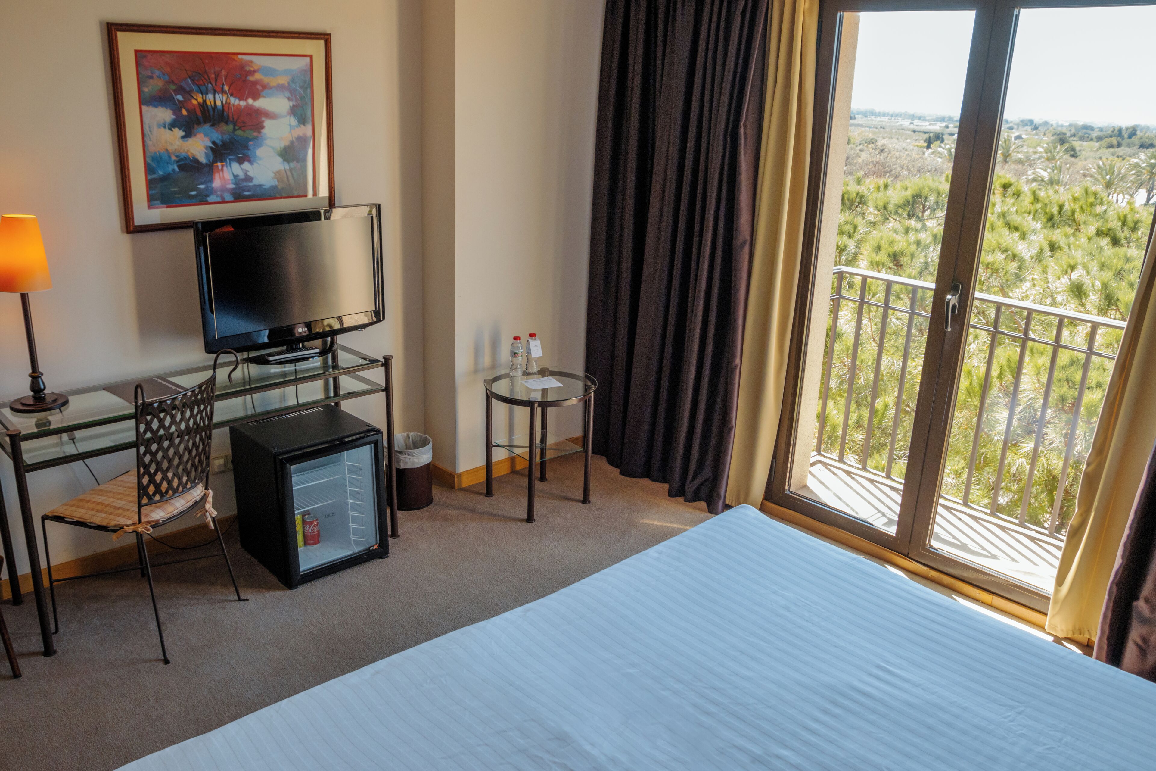 A hotel room with a bed, a desk with a television, a small refrigerator, a chair, a trash can, and a view of the outdoors through a balcony.