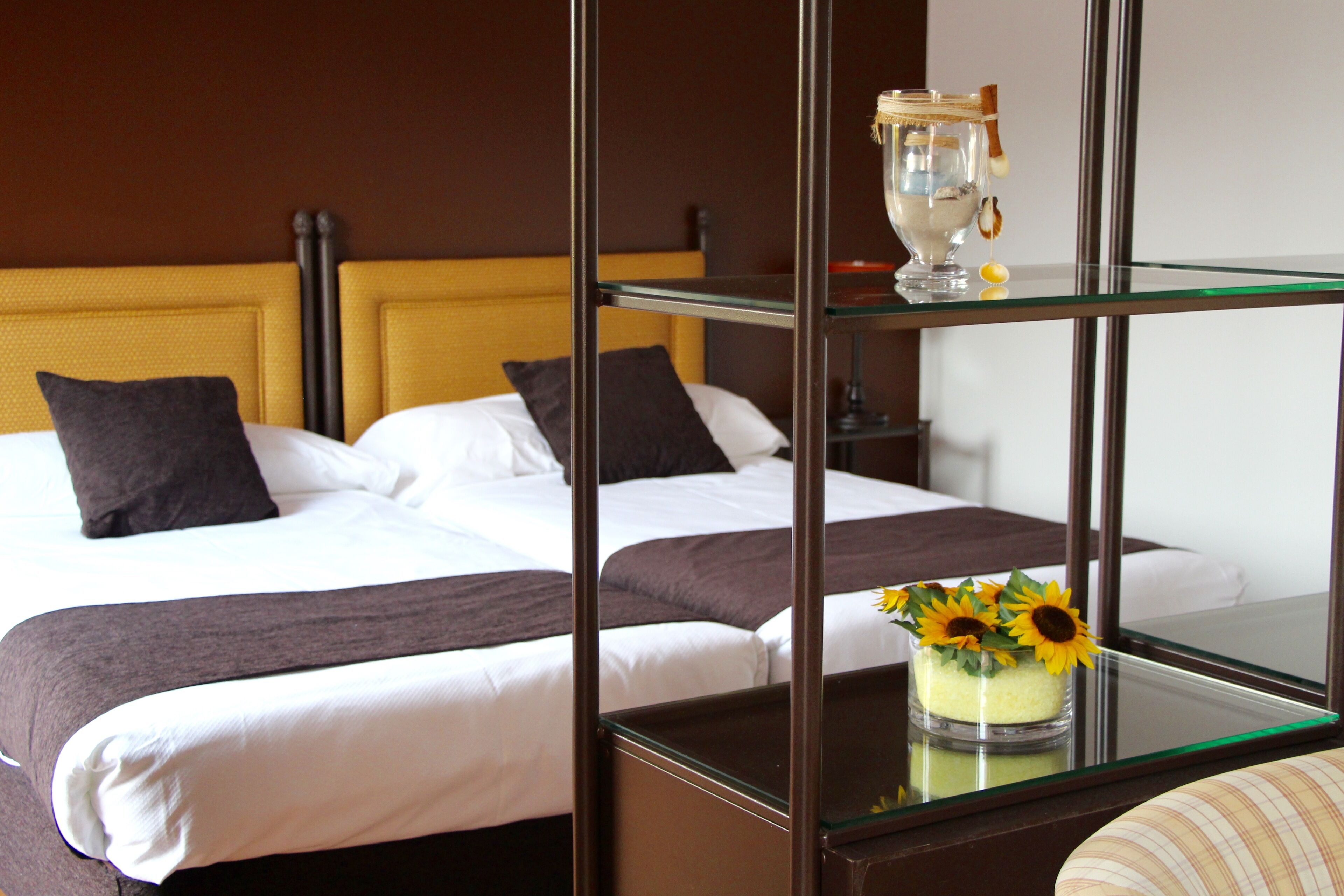 Habitación de hotel con dos camas, una mesita de vidrio con un jarrón de girasoles y un espejo enmarco.