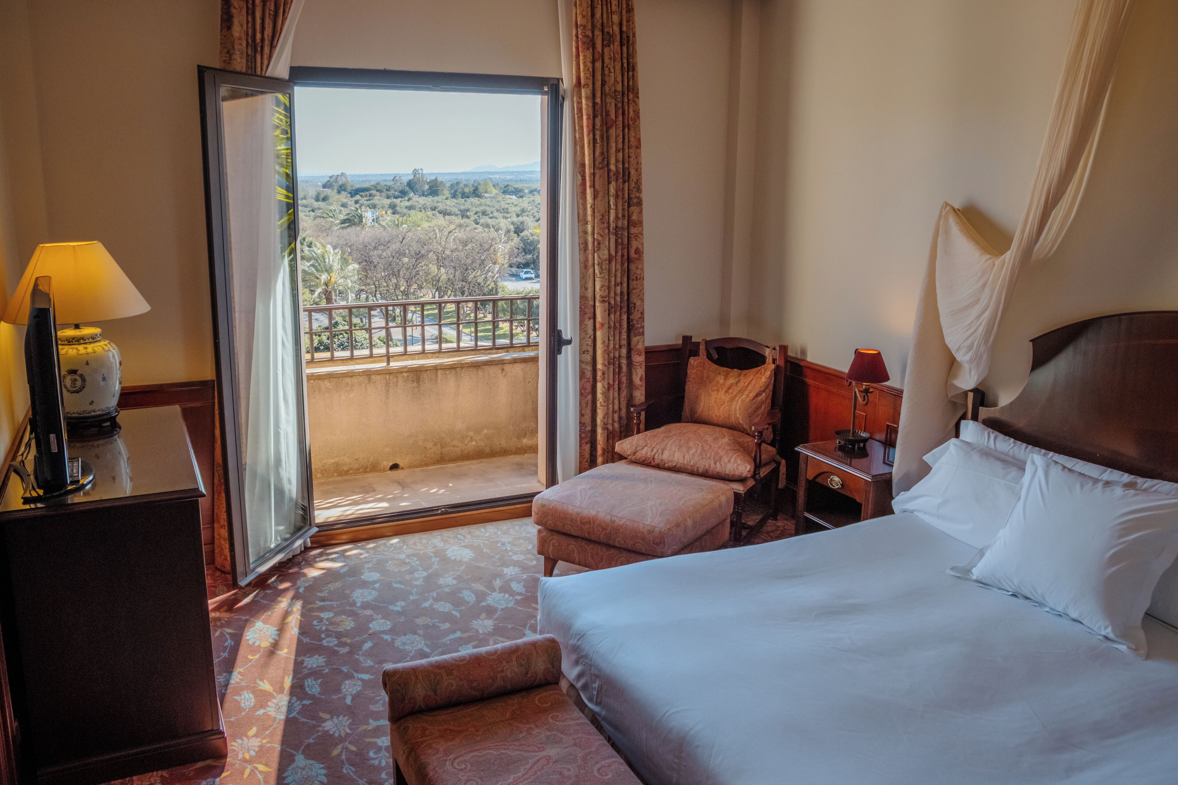 Una habitación de hotel con cama, una silla, un escritorio con lámpara y vistas al exterior a través de una puerta corrediza.