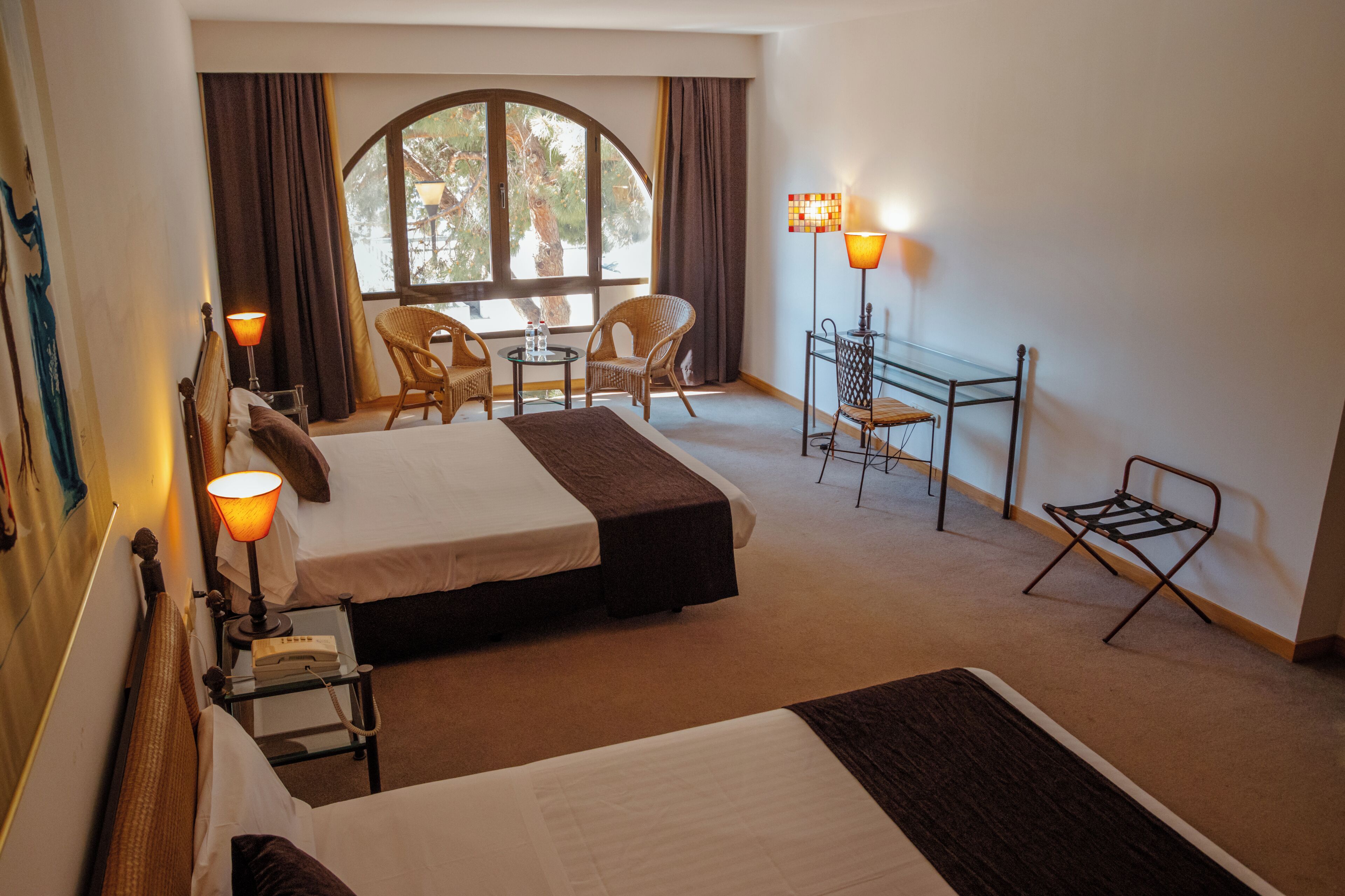 A hotel room with two beds, a small table, two chairs, a lamp, and a window with curtains.