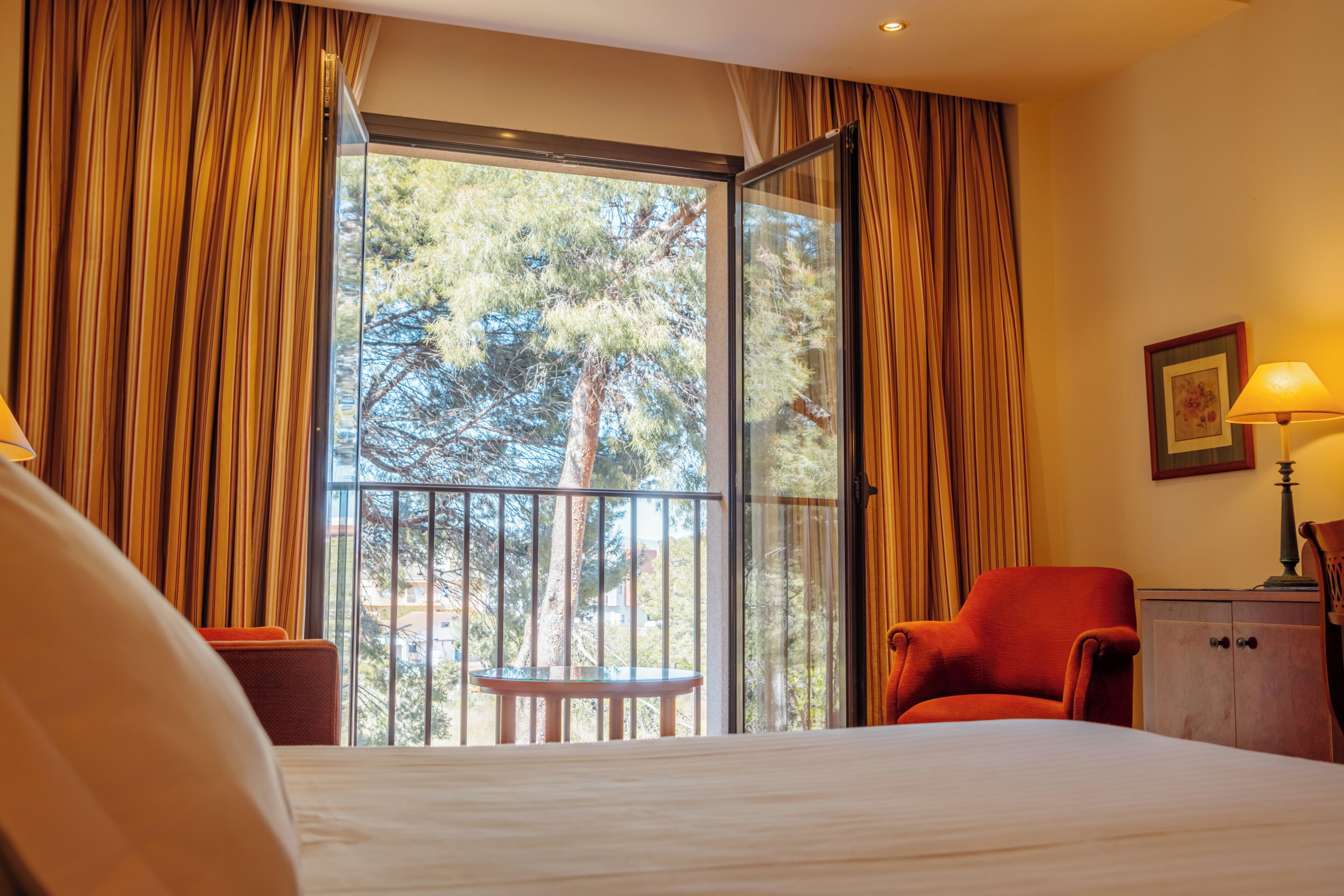 A hotel room with a large bed, a red armchair, a small table, and a balcony with curtains.