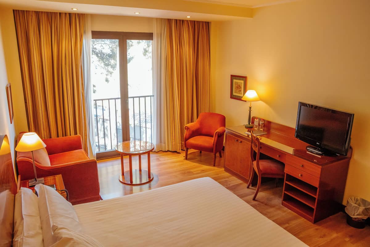 A hotel room with a bed, a desk with a television, a chair, a table, and a window with curtains.