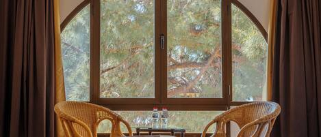 A room with a bed, two wicker chairs, a small table with water bottles, and a large arched window overlooking trees.