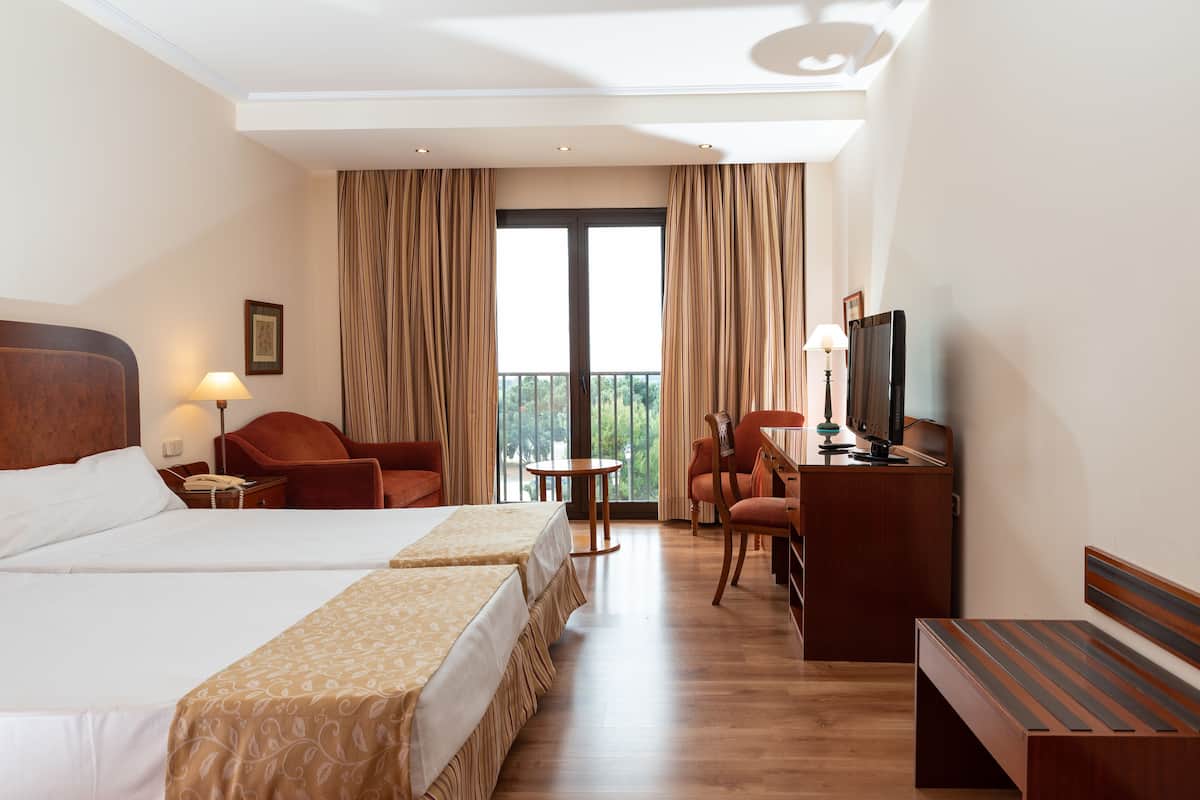 A hotel room with two beds, a desk, a chair, a TV, and a balcony with curtains.