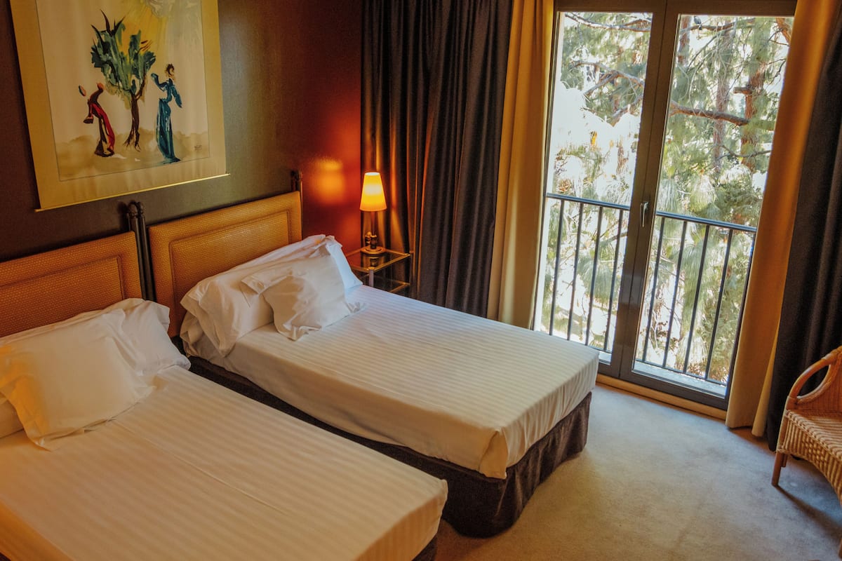A hotel room with two beds, a nightstand with a lamp, a painting on the wall, and a balcony with a view of trees.