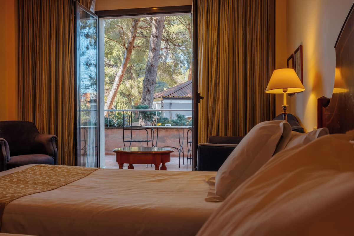 A hotel room with a bed, a chair, a table, and a view of trees and a building through a glass door.