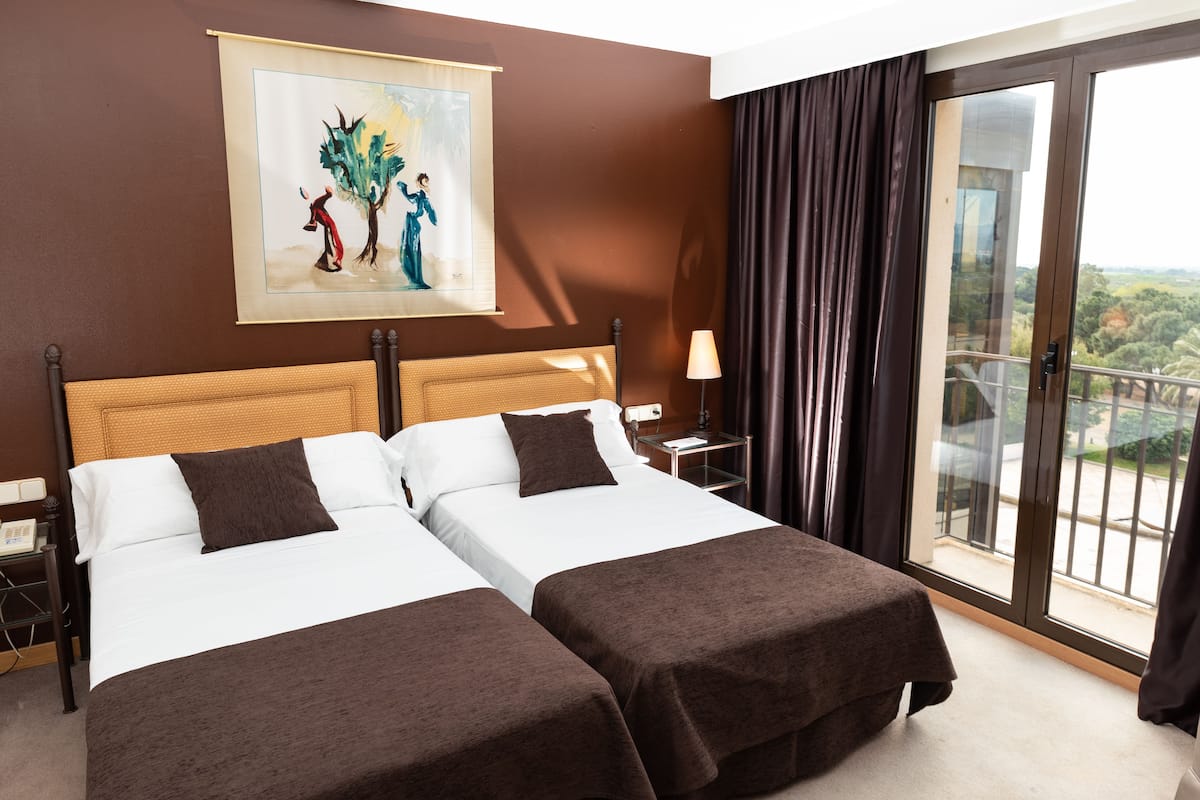 A hotel room with two beds, a painting on the wall, a nightstand, a lamp, and a balcony with a view.