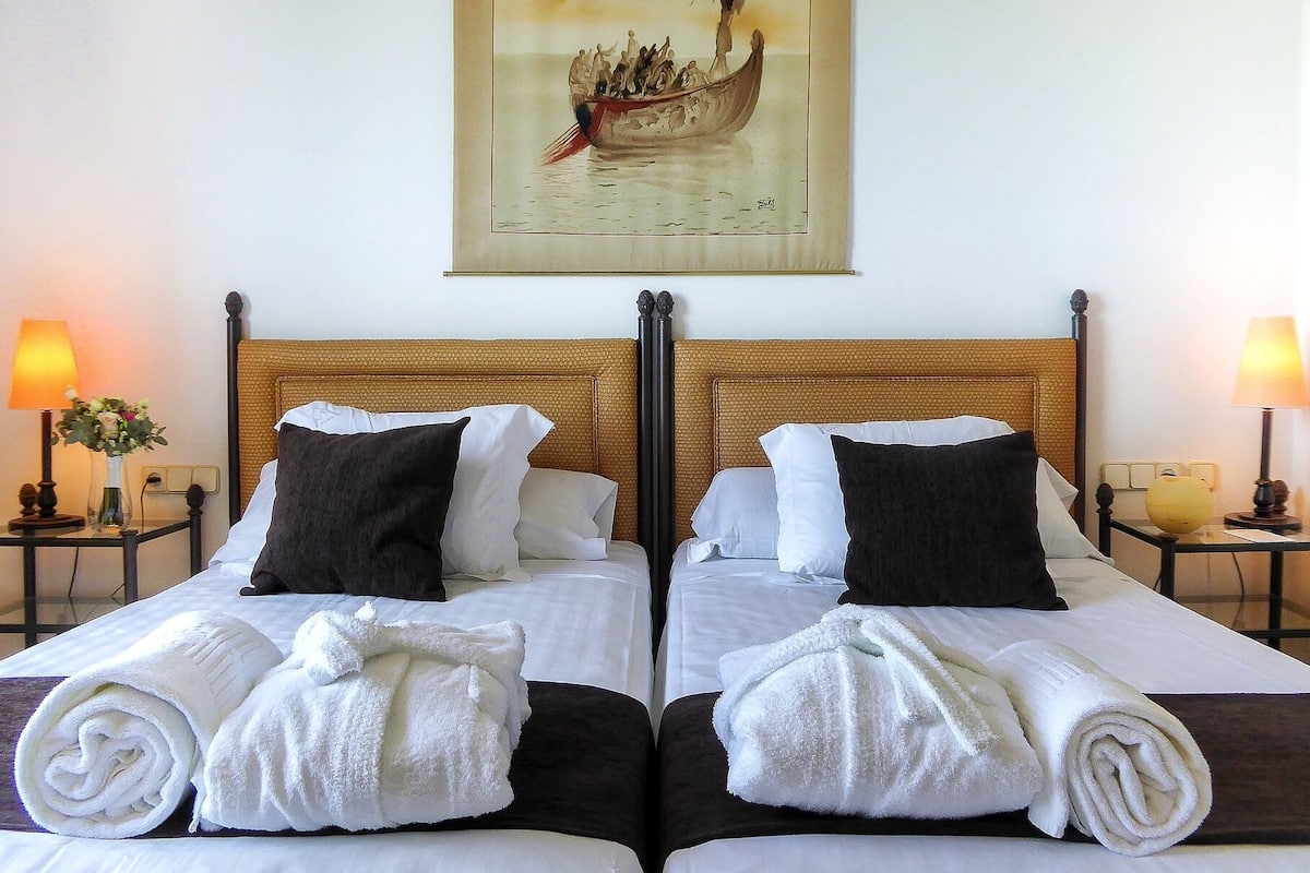 A double bed with white linens, two black pillows, and rolled white towels. There are two bedside tables with lamps and a painting of a boat on the wall.
