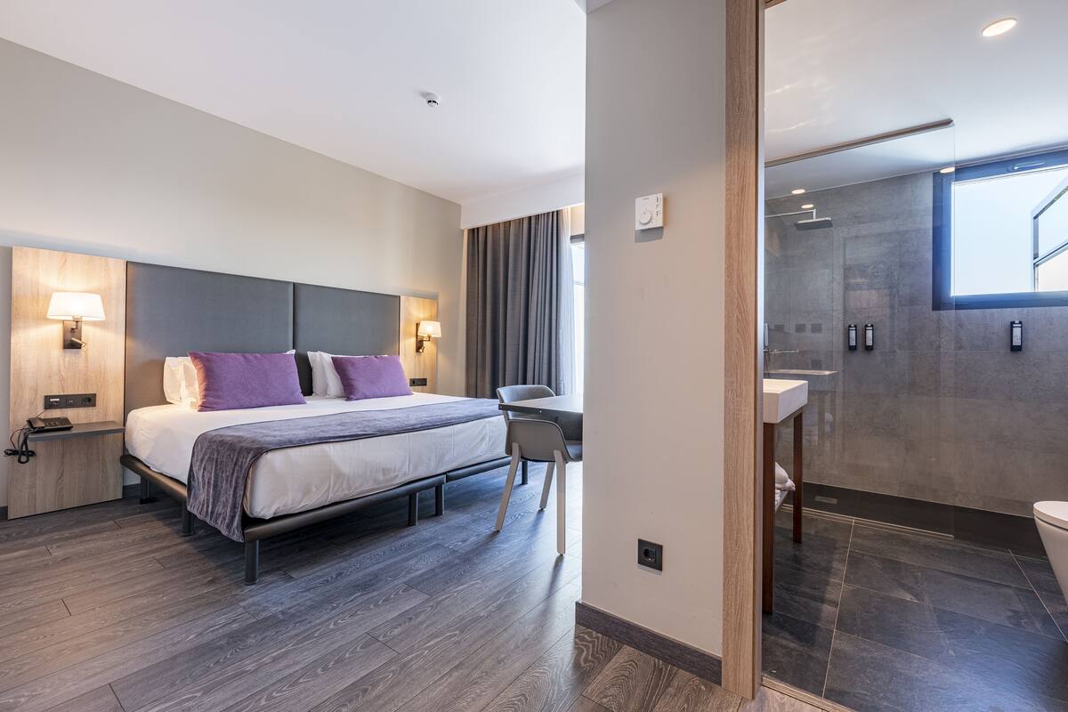 A modern hotel room with a bed, bedside tables, a desk, and a shower area.