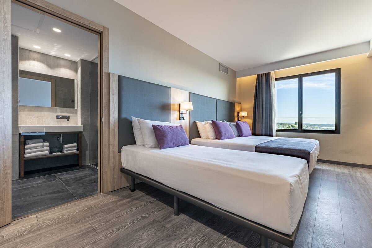 A hotel room with two beds, a bathroom with a vanity, and a large window offering a view.