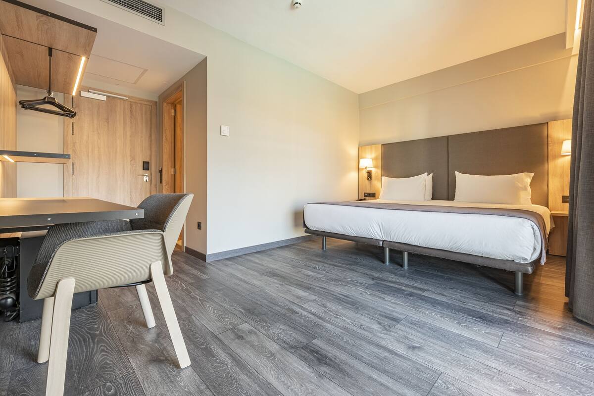 A modern hotel room with a bed, bedside table, chair, and a desk with a chair.