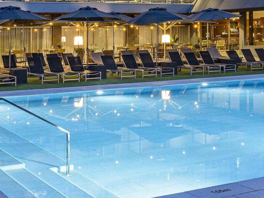 A modern hotel pool area with lounge chairs and umbrellas, a glass barrier, and a well-lit interior.