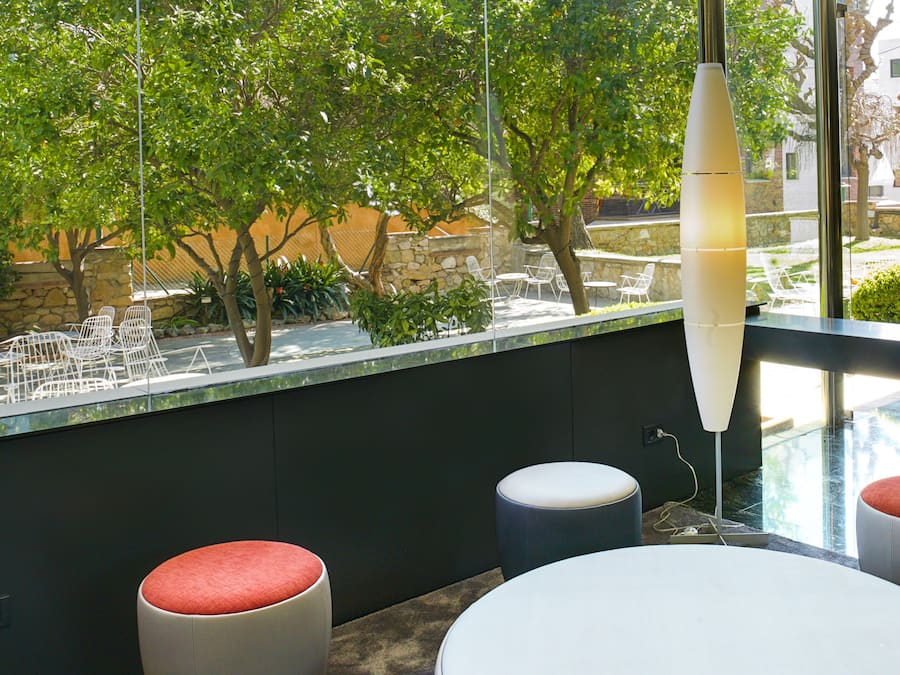 Un área de estar moderna al aire libre con sillas blancas y una mesa redonda, una lámpara de pie alta y vistas a una calle arbolada.