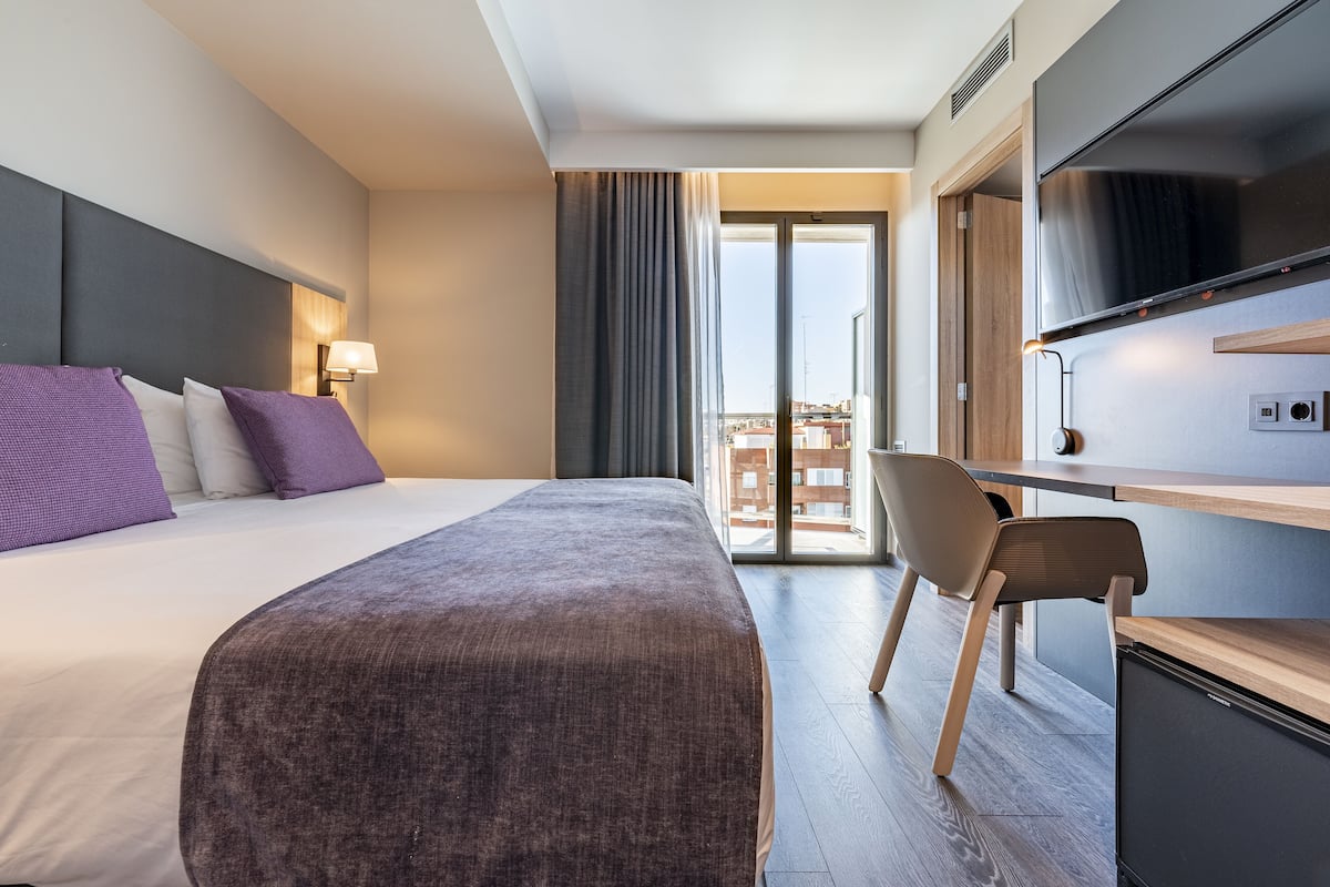 A hotel room with a bed, a desk, a chair, a TV, and a view of the city.