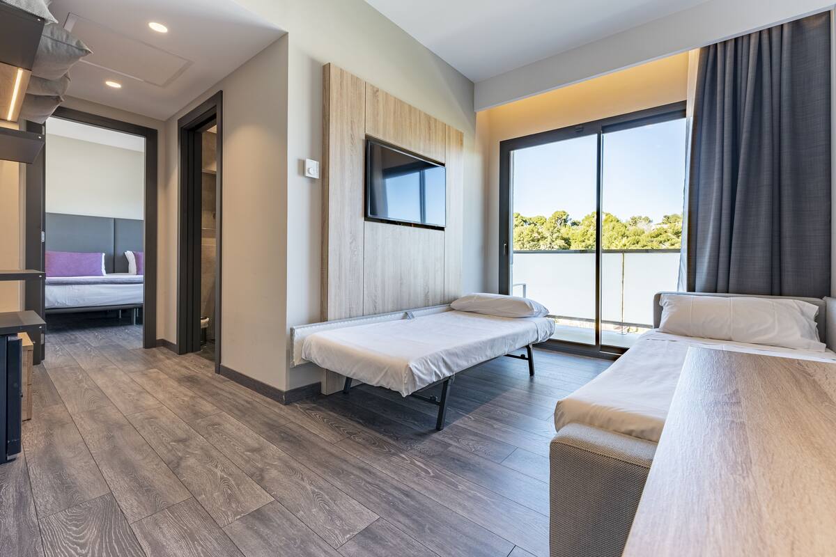 A modern hotel room with a bed, a TV mounted on the wall, and a balcony with a view of trees.