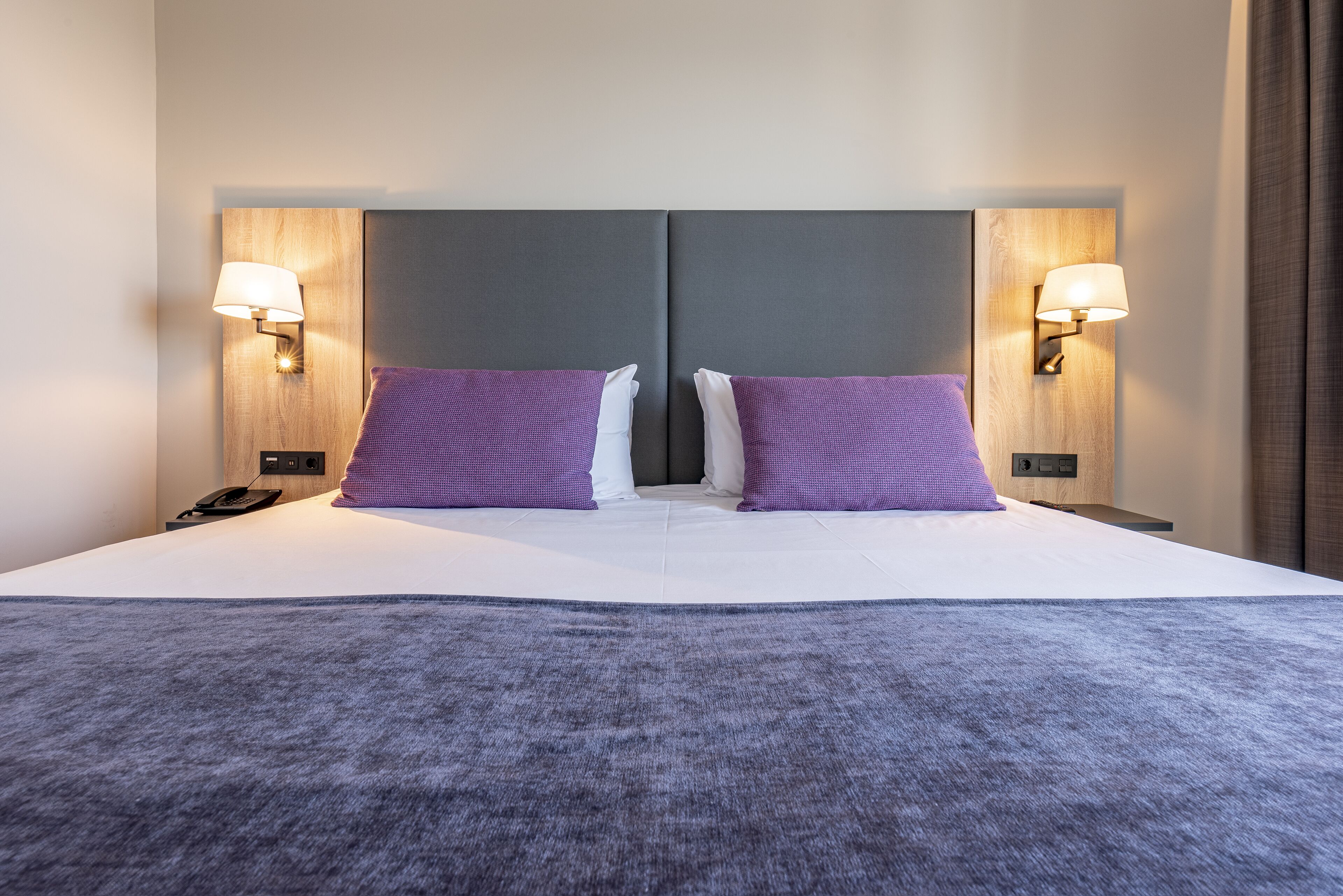 Habitación de hotel con una cama grande, dos almohadas moradas, un cabecero gris y lámparas fijadas a la pared.