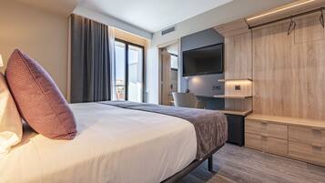 A modern hotel room with a large bed, a flat-screen TV mounted on the wall, a desk with a chair, and a view of the city through the window.