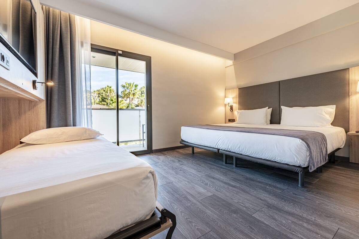 A hotel room with a bed, a second bed, a nightstand, a lamp, and a sliding glass door leading to a balcony.