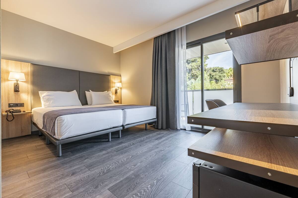 A modern hotel room with a bed, bedside lamps, a desk, and a large window with curtains.
