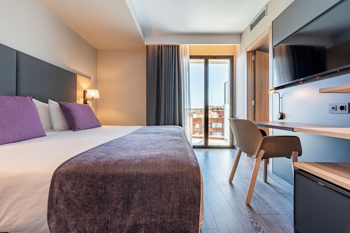 A modern hotel room with a large bed, a desk with a chair, a flat-screen TV, and a sliding glass door to the balcony.