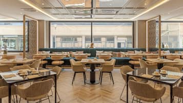Un comedor moderno con varias mesas y sillas, una zona de estar central con sofás, y grandes ventanales que ofrecen vistas de la ciudad.